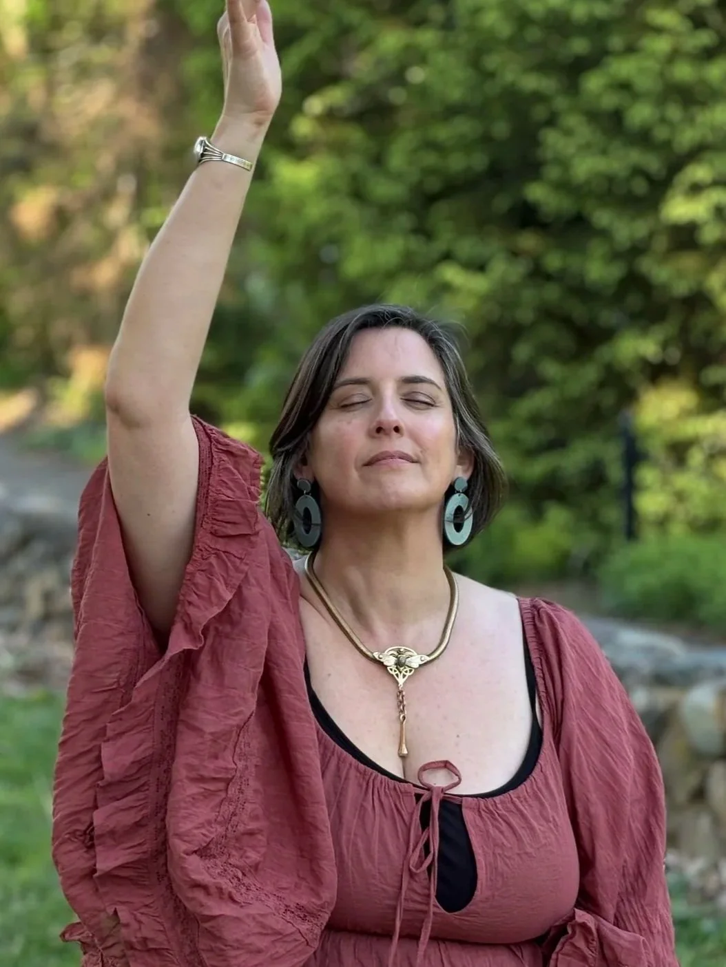 A woman with short dark hair and closed eyes standing outdoors with a relaxed expression, her right arm raised, wearing a reddish-brown loose blouse, large earrings, a necklace, and a silver bracelet, with a background of green trees and soft natural light.