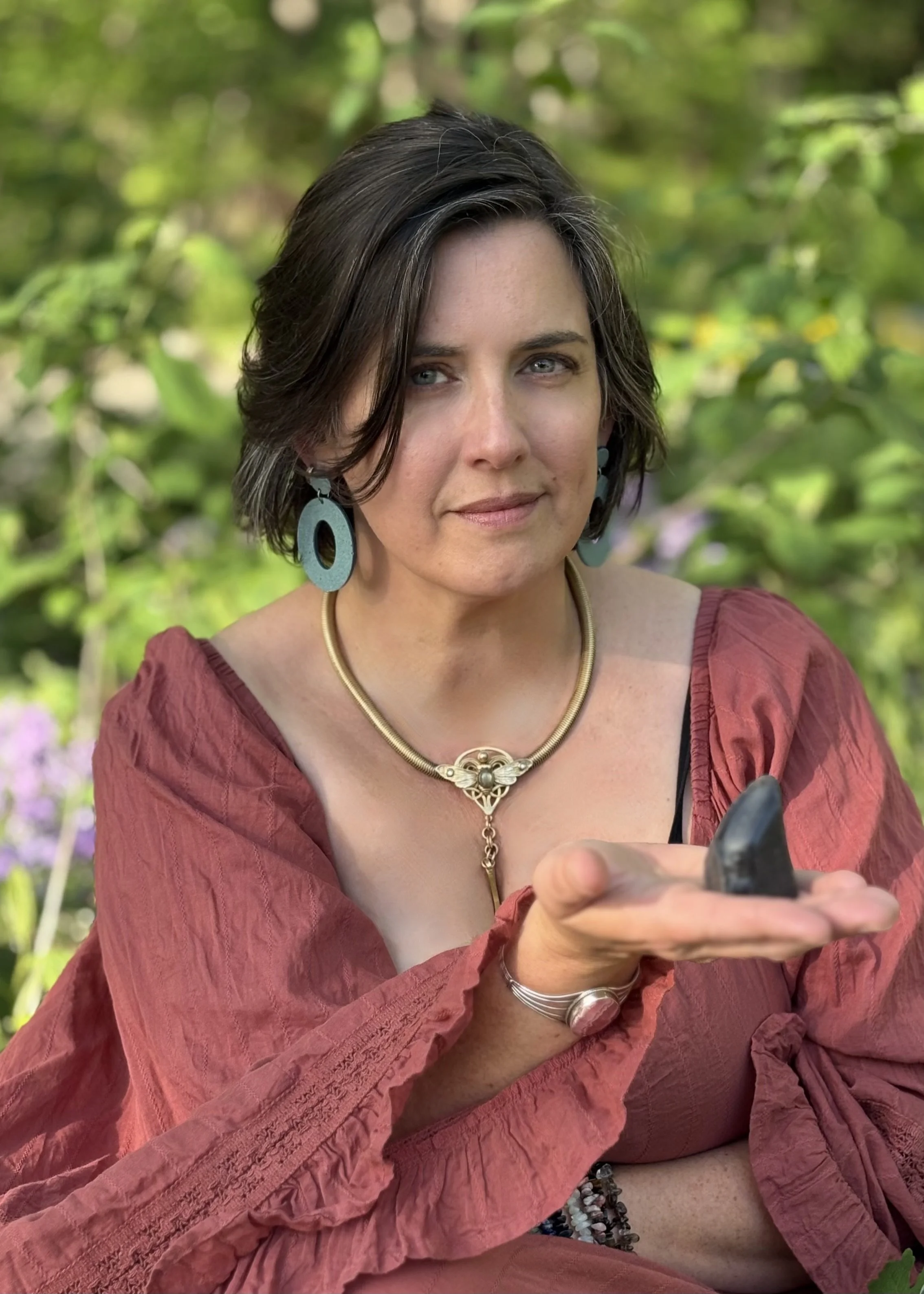 A woman with short dark hair and blue eyes wearing large blue earrings, a gold necklace, a gold chain with a pendant, and a silver bracelet, sitting outdoors in a green leafy setting while looking at her smartphone.