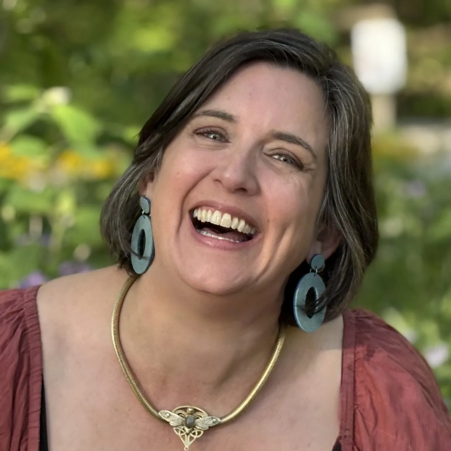 Close-up of a smiling woman with short dark hair, wearing large blue earrings, a gold necklace, and a red top, outdoors with a blurred green background.