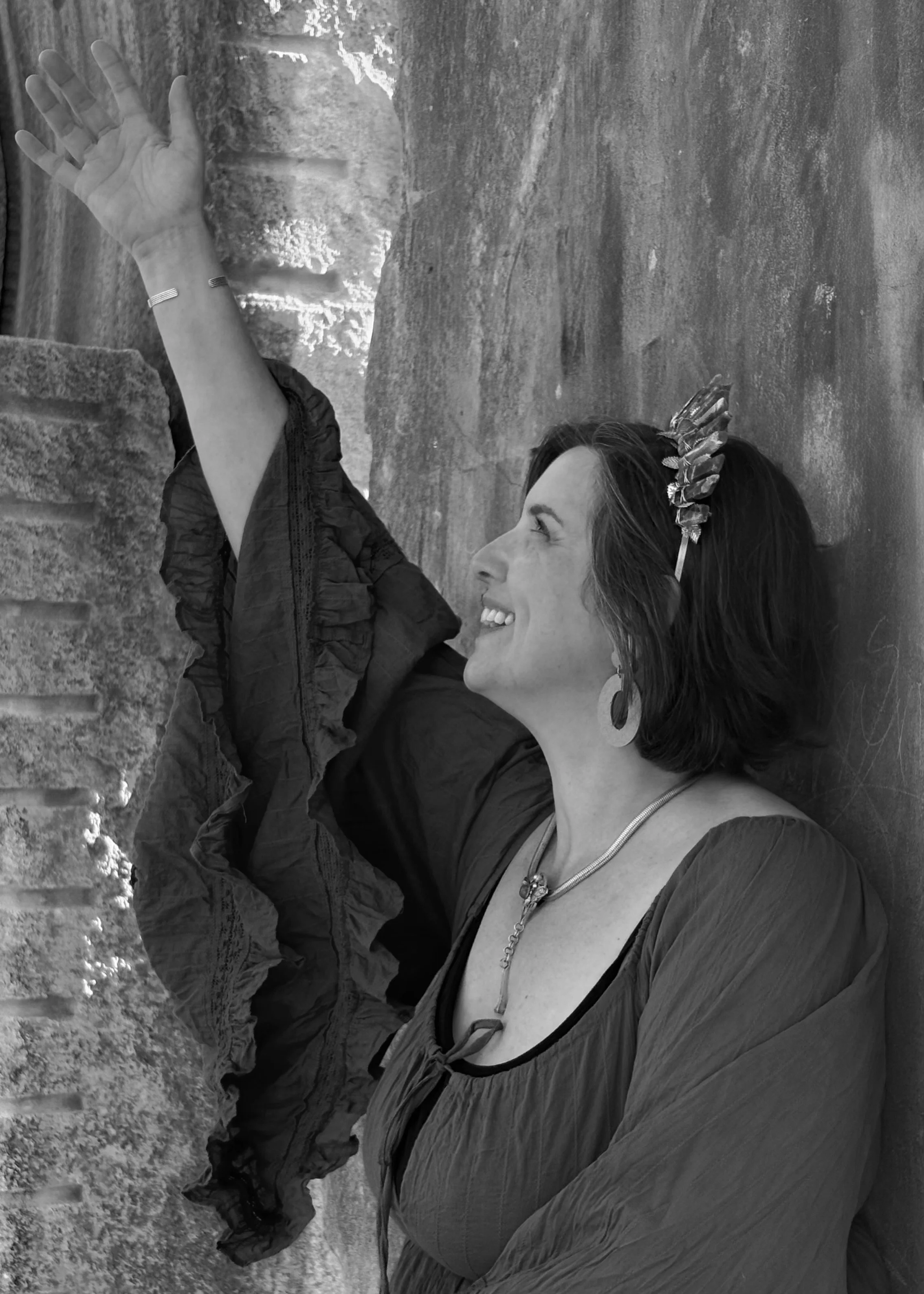 A woman lying on her side on the ground, smiling and looking up at a rock face, with her hand touching the rocks. She is wearing earrings, a necklace, and a dark blouse with a ruffled sleeve. Her hair is short and styled, and she has a decorative headband.