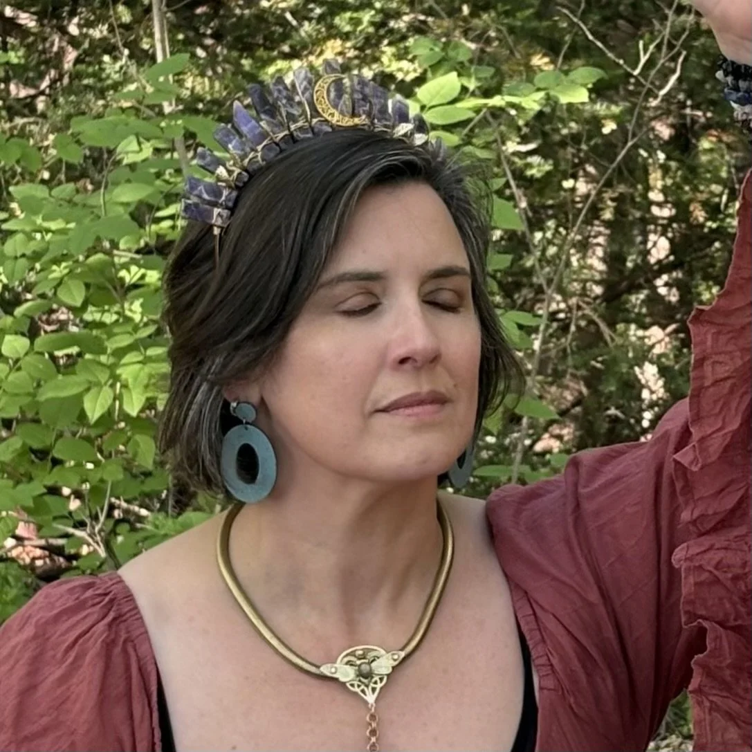 A woman with closed eyes wearing large blue earrings, a gold necklace, and a decorative headpiece, standing outdoors surrounded by green foliage.