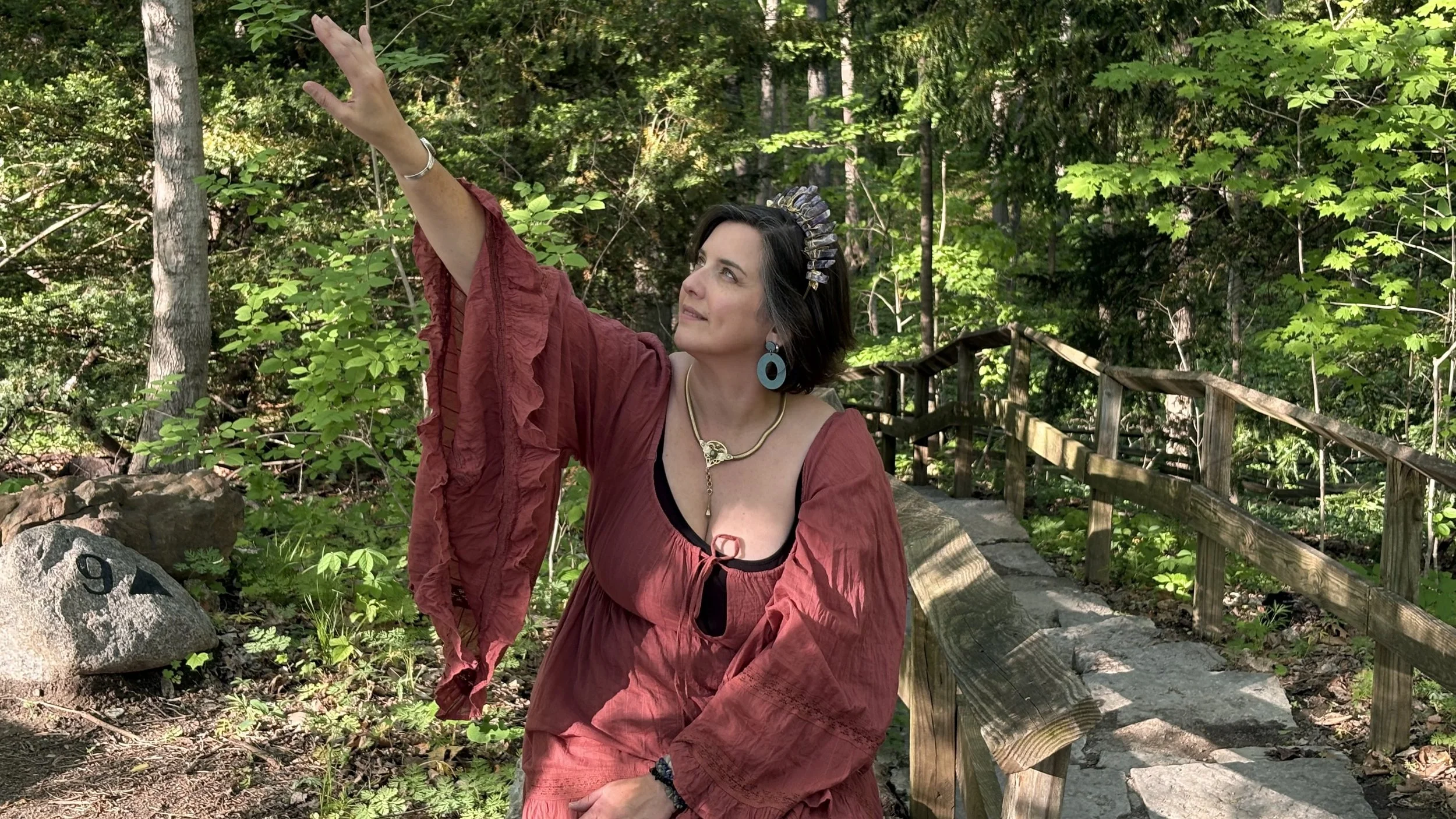 A woman with dark hair, wearing a rust-colored dress, is standing on a forest trail with a wooden railing. She is reaching up with her right arm, and is accessorized with large blue earrings, a necklace, and a headpiece. The background consists of trees and greenery.