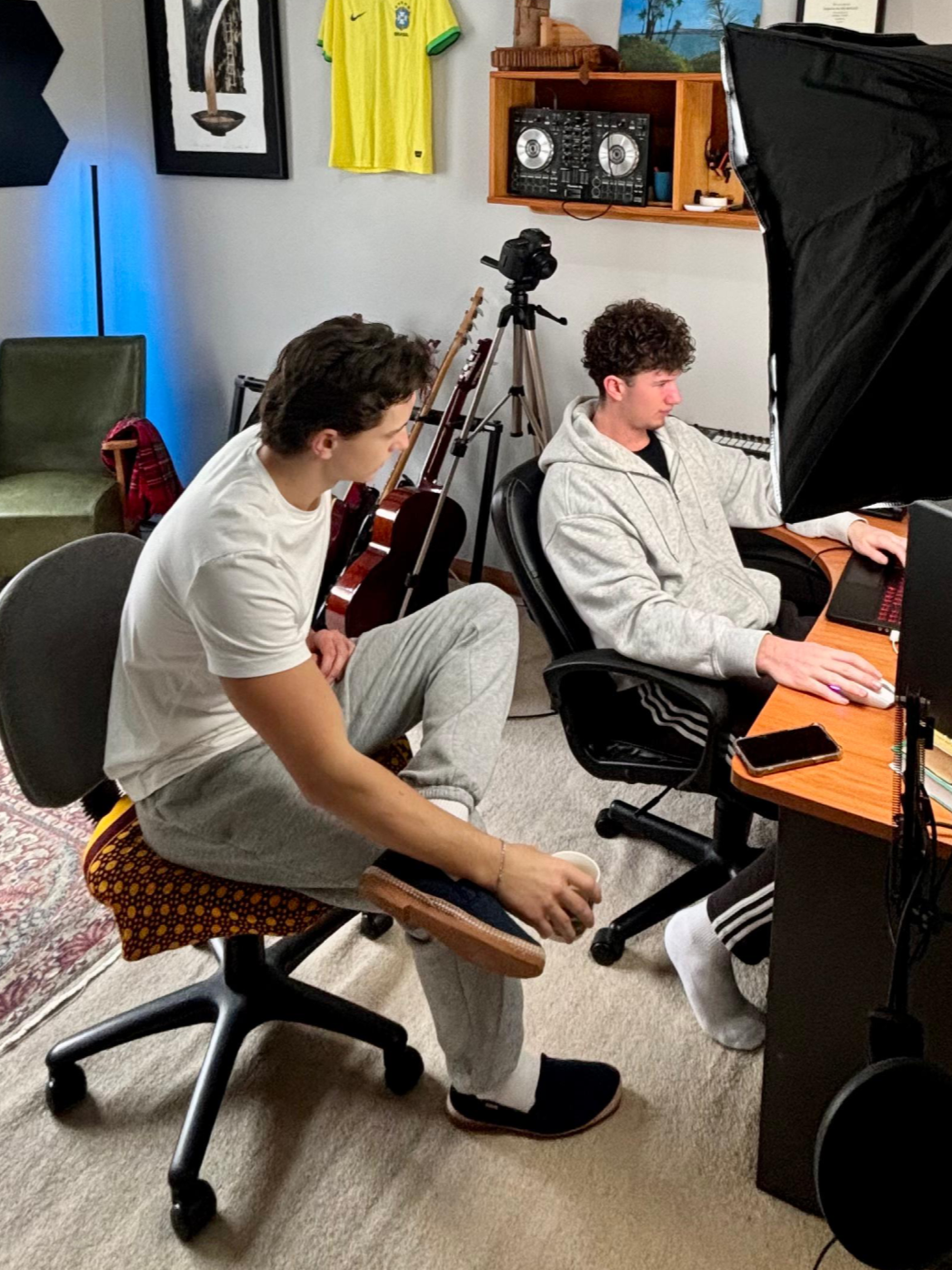 Two young men in a home recording studio with musical equipment. One sits at a desk with a computer, the other sits on a chair with a mug and a footrest.