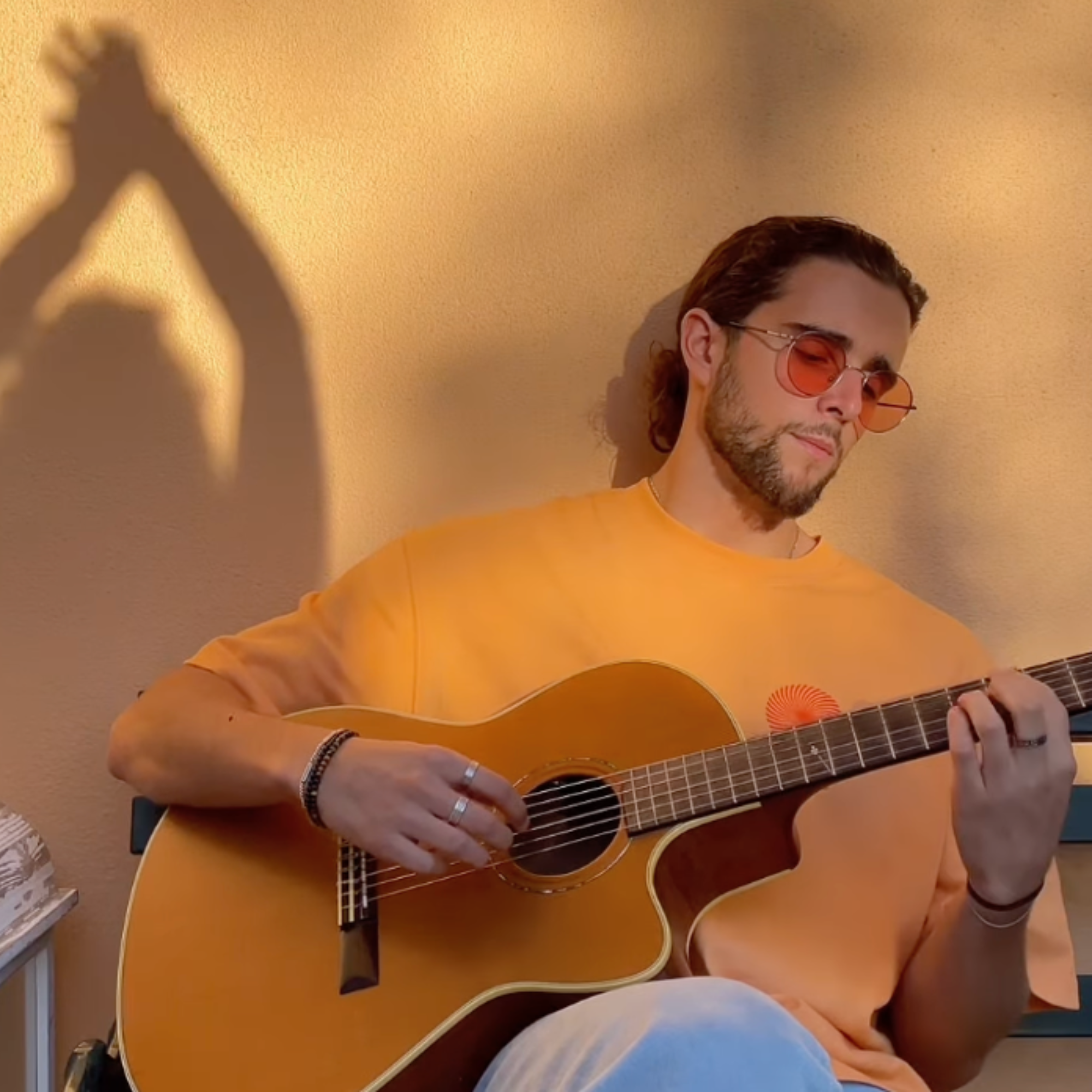 A young man with sunglasses playing an acoustic guitar against a beige wall, casting a shadow on the wall.