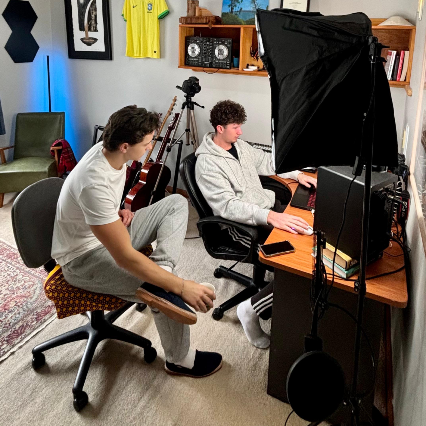 Two young men in a music studio setup; one is sitting at a computer wearing a gray hoodie, and the other kneeling on a chair with a cup in hand. Music equipment, guitars, and books are visible in the background.