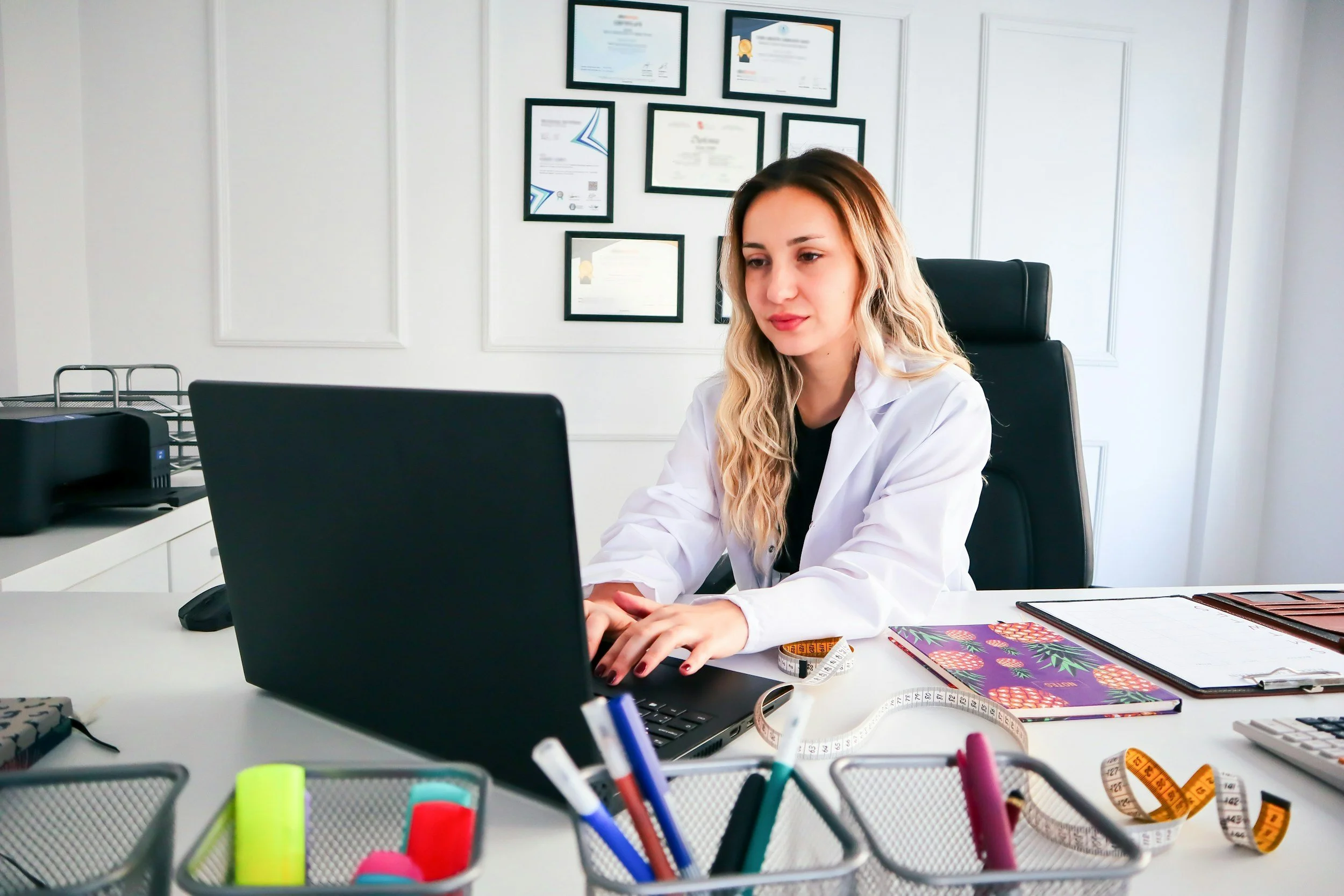 A woman in a white coat working on a laptop at her desk in an office, with framed certificates on the wall behind her.