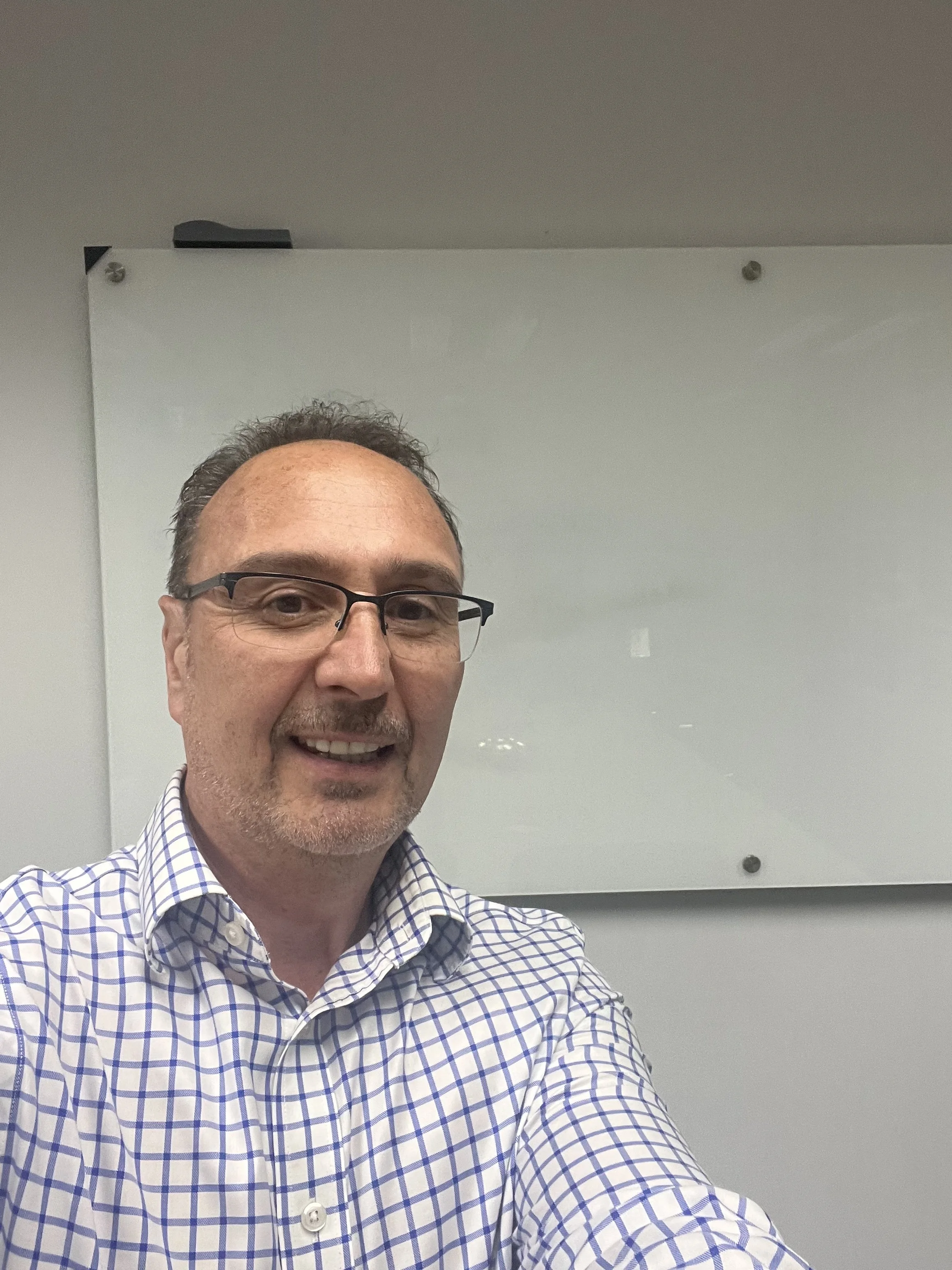 A middle-aged man with glasses and a checkered dress shirt taking a selfie in front of a whiteboard in an office or conference room.