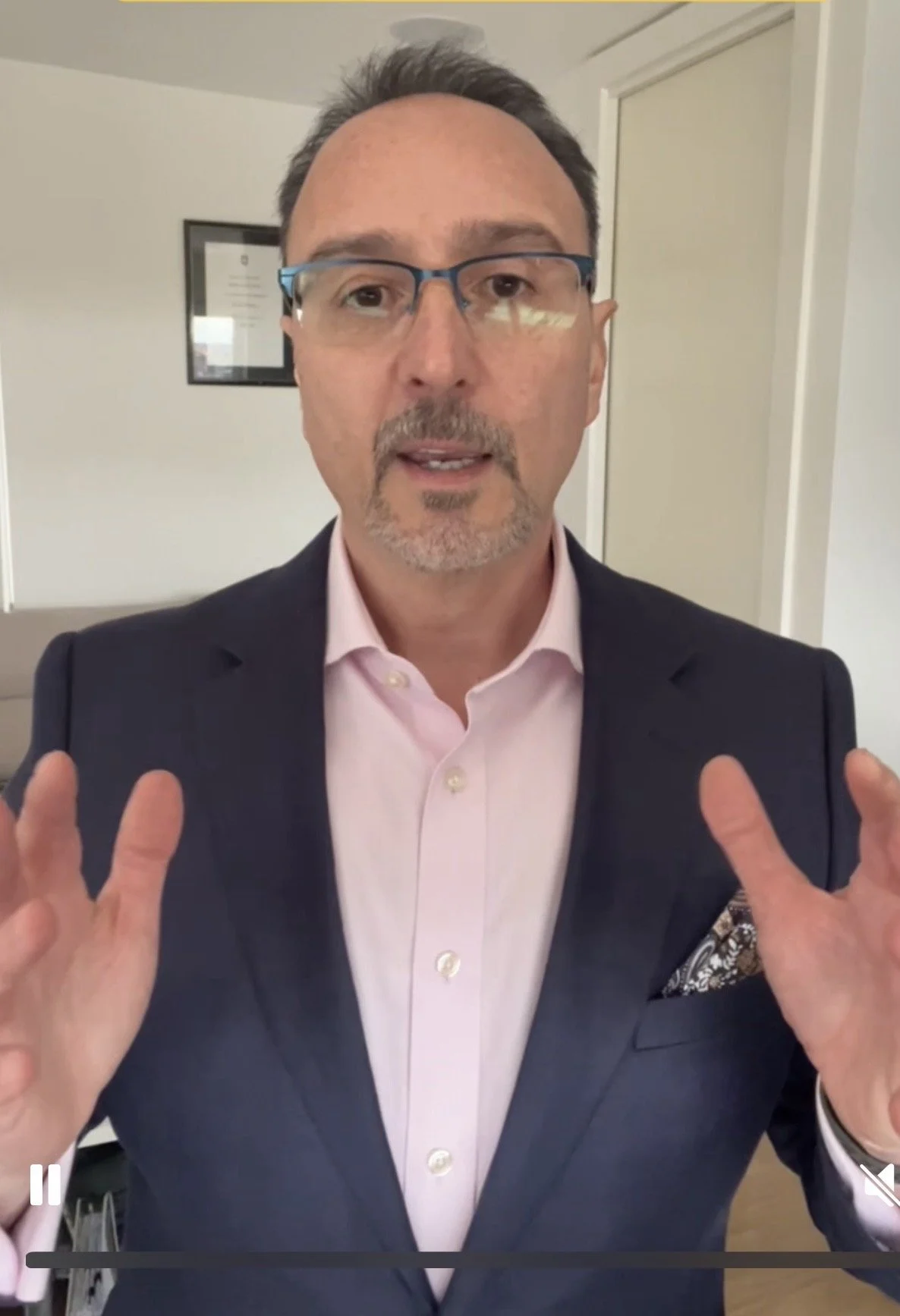 A middle-aged man with glasses, wearing a pink dress shirt and dark blazer, is speaking and gesturing with both hands.