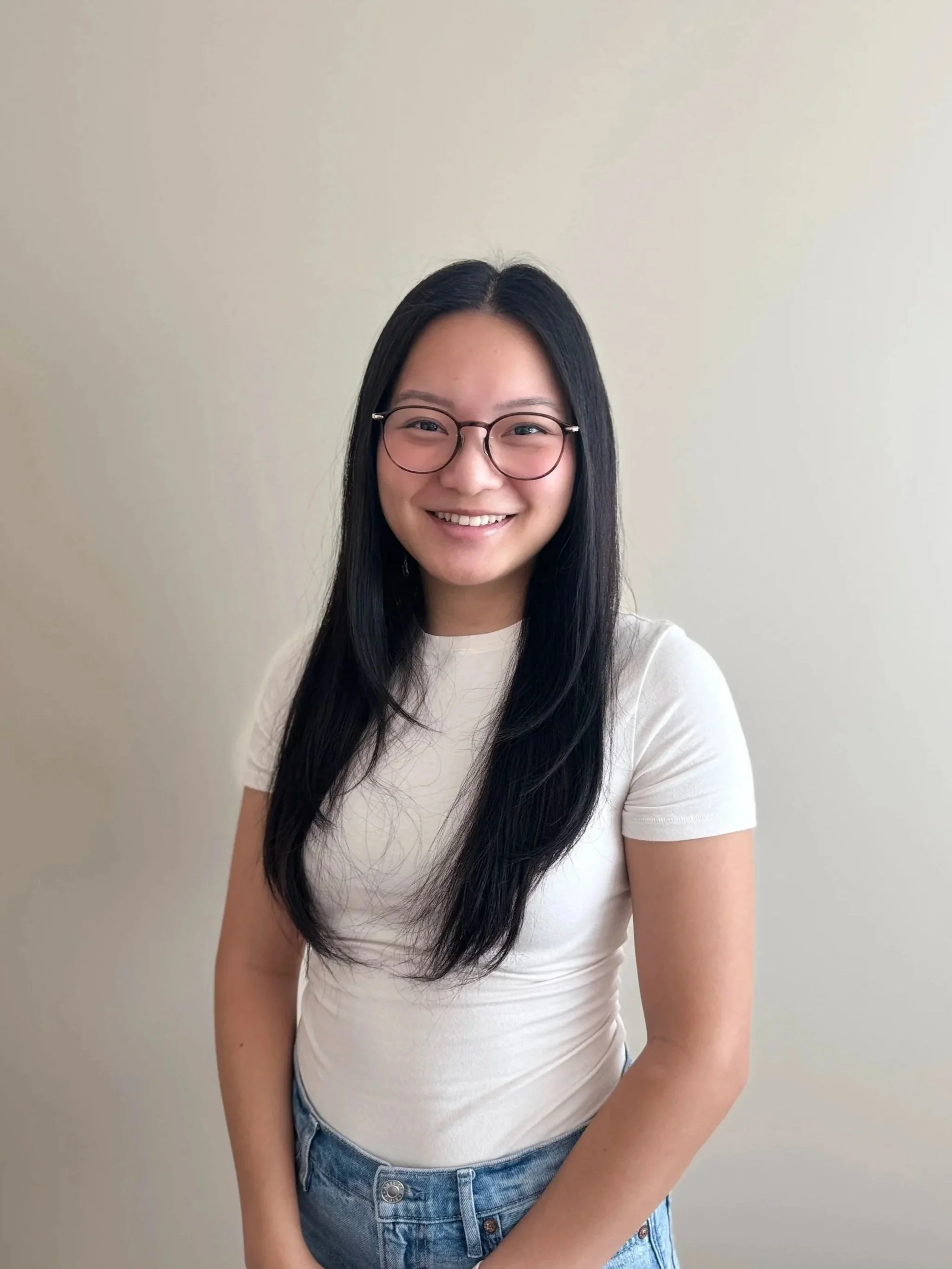 A girl with long black hair and glasses smiles warmly. She is wearing a white t-shirt and blue jeans and is standing against a plain beige wall.