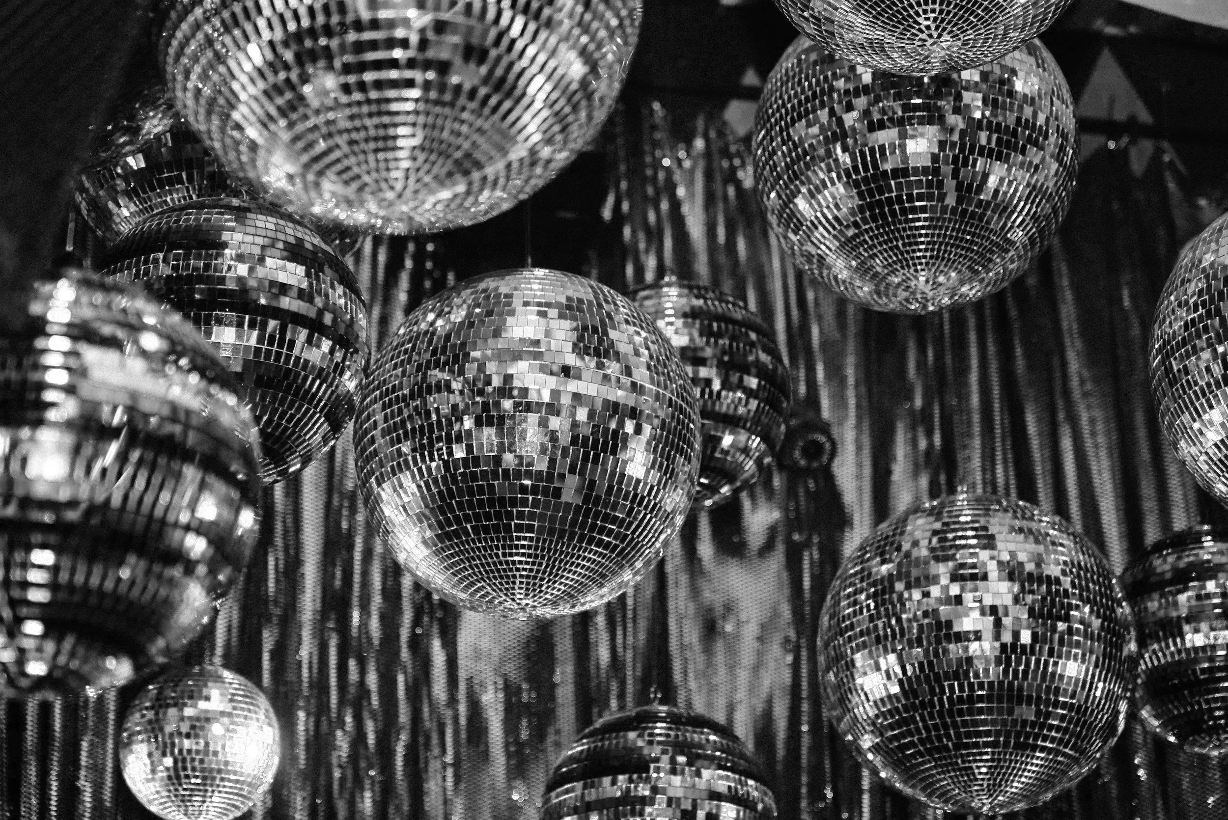 Black and white image of multiple disco balls hanging, reflecting light off mirrored tiles. The scene evokes a retro, festive atmosphere.