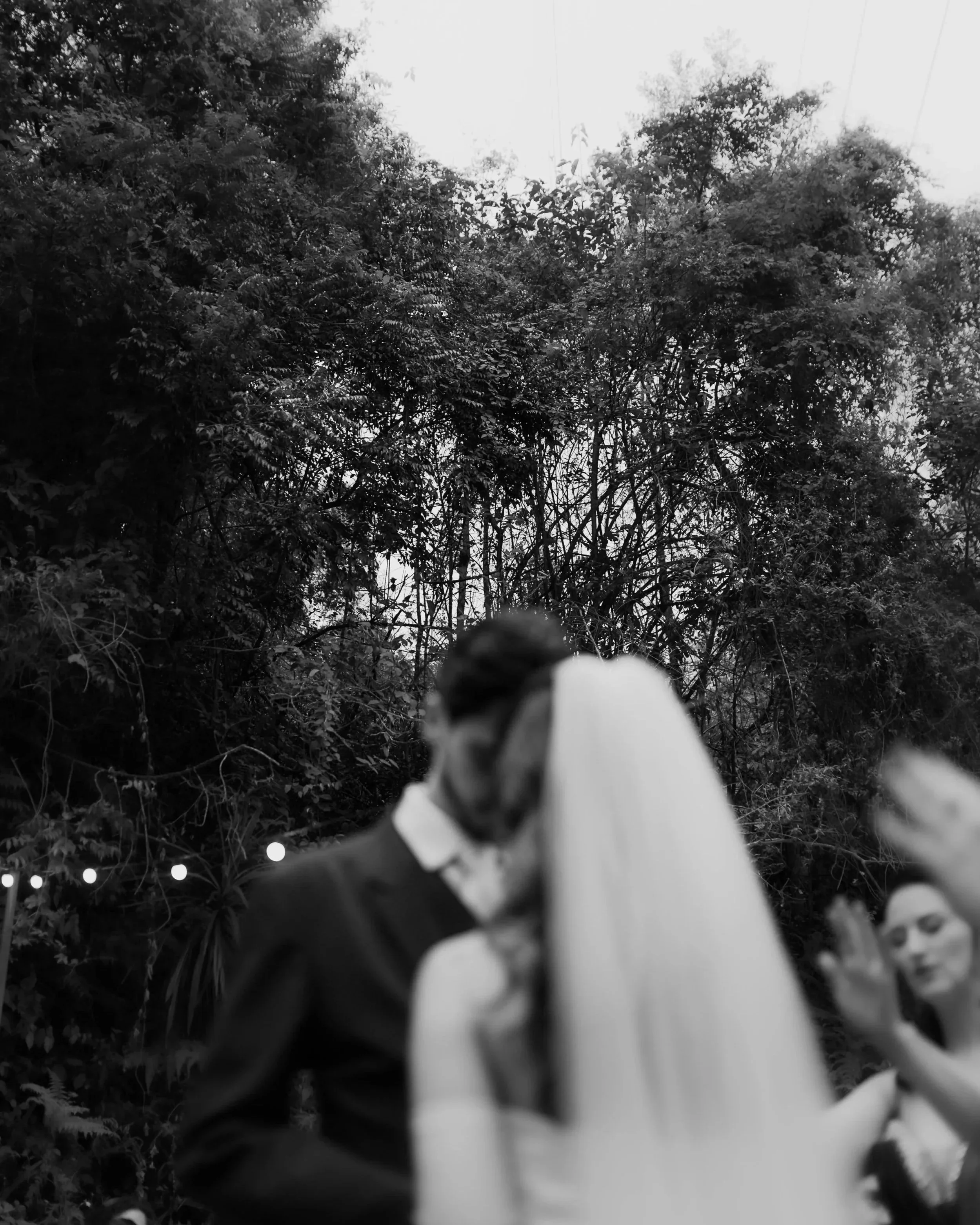 A blurred black-and-white photo of a wedding couple embracing outdoors, with trees in the background. A guest appears clapping, adding to the joyful ambiance.