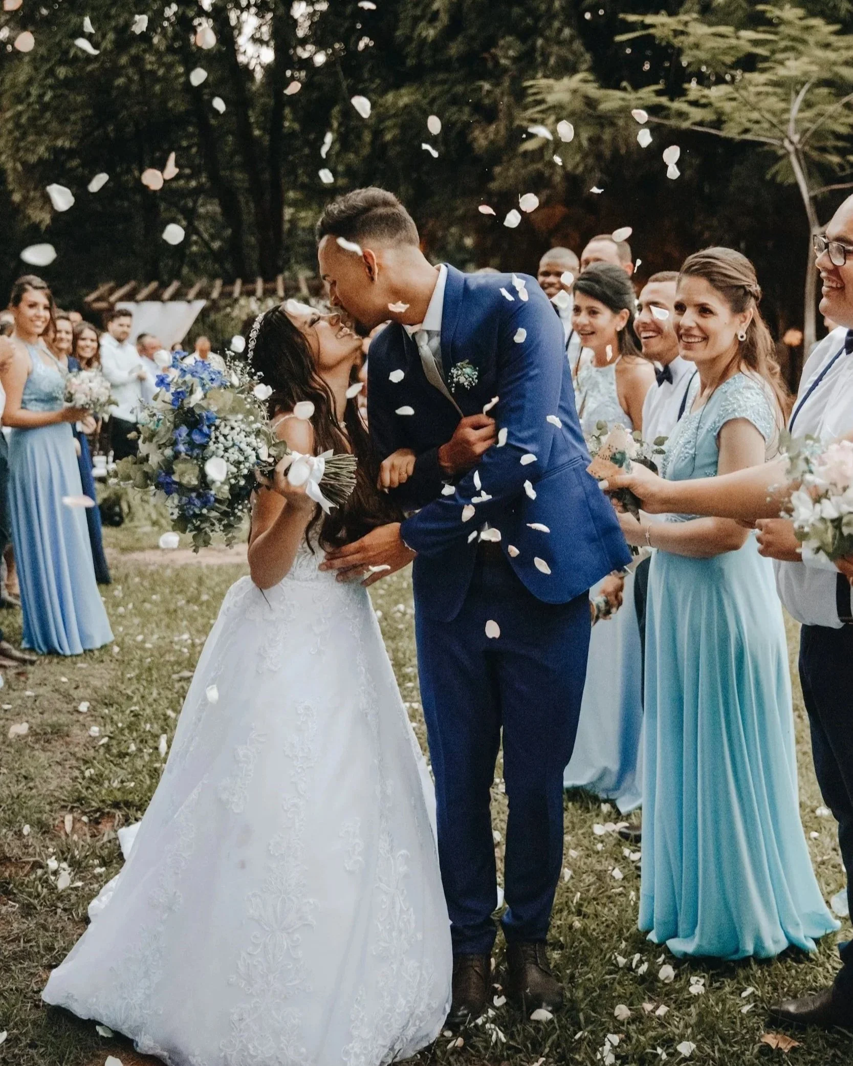 A bride and groom kiss outdoors, surrounded by smiling bridesmaids in blue. Flower petals fall around them, creating a joyful and romantic scene.