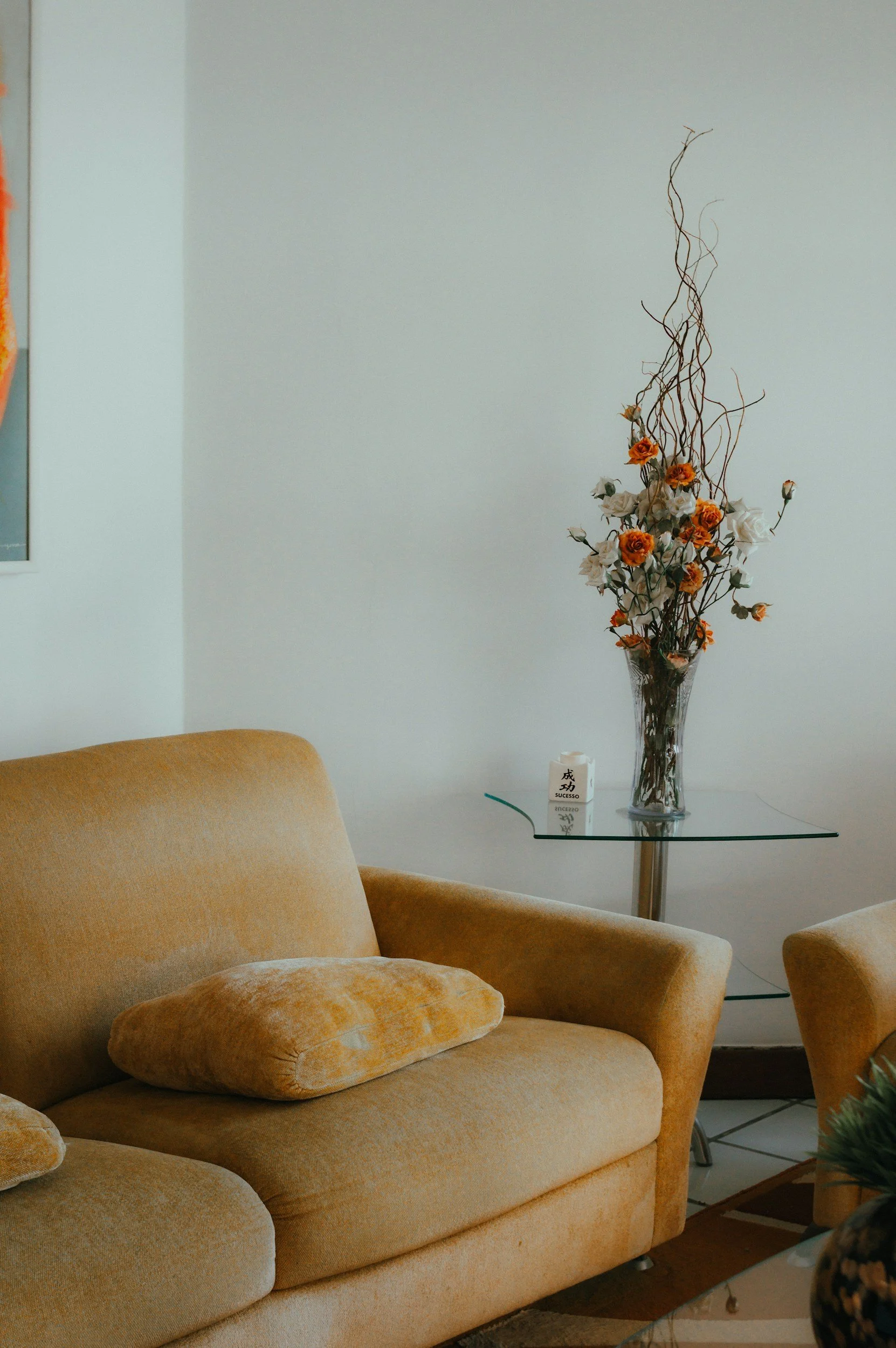 A yellowish-beige upholstered sofa with a matching pillow, in a room with white walls. Behind the sofa, a glass side table holds a tall glass vase with orange and white flowers and curly twigs. Part of a framed picture is visible on the left wall.