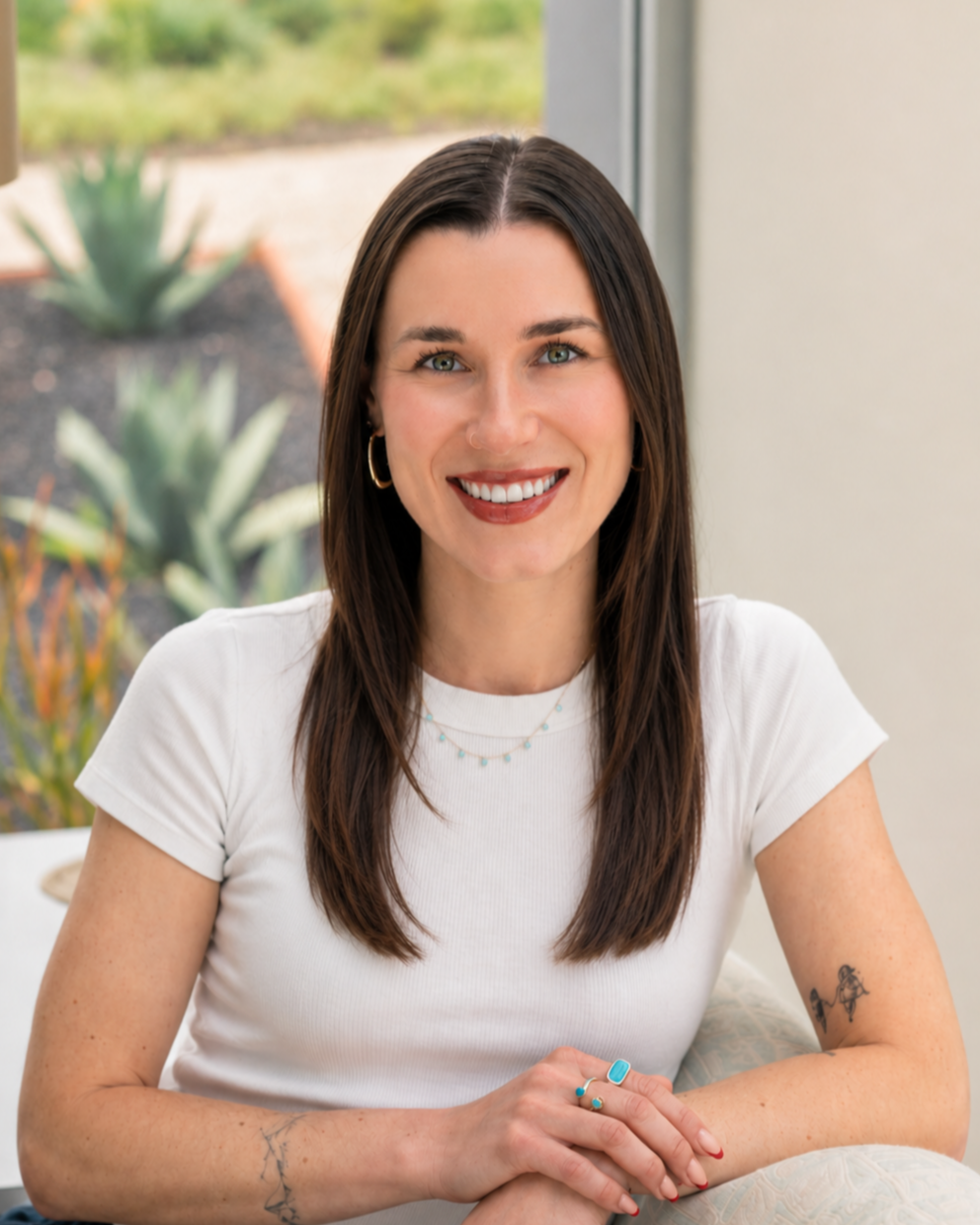 A woman with dark brown hair, wearing a white T-shirt, gold hoop earrings, and layered necklaces, smiling while sitting indoors near a window with a view of succulents outside.