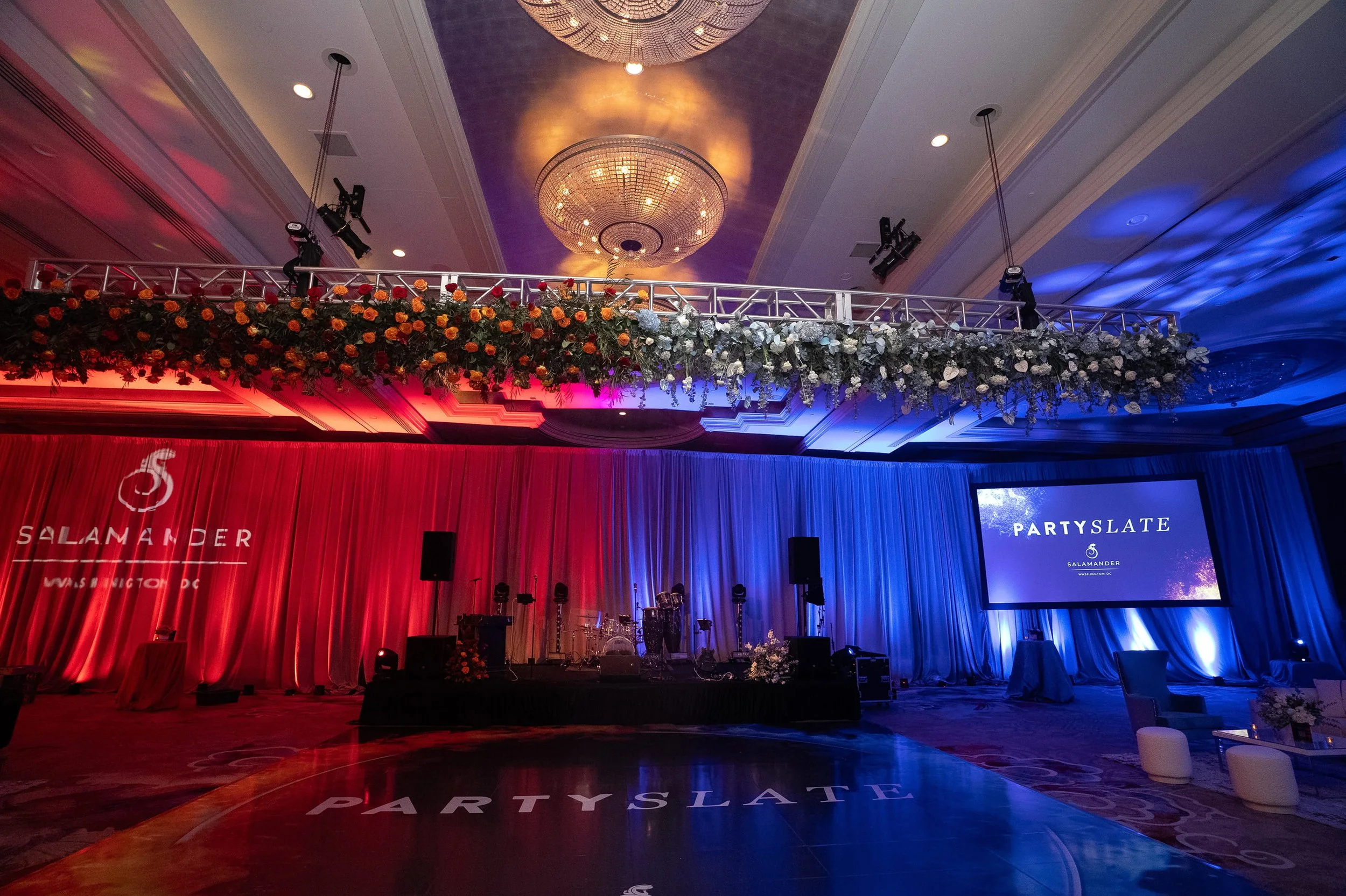 Ballroom event setup with floral truss overhead, red and blue uplighting, and projection screen by RDAV