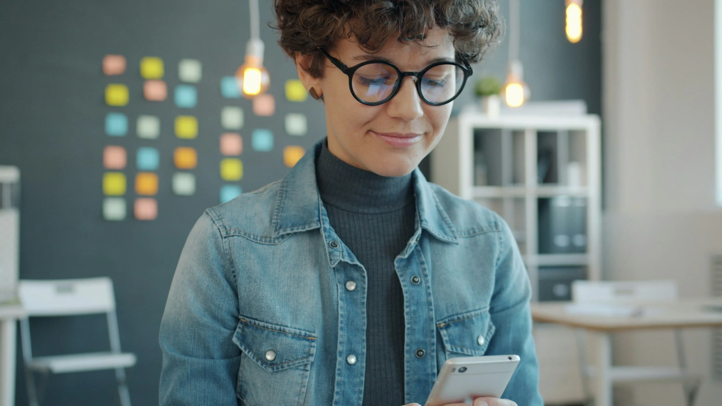 A young woman with short curly hair, glasses, and a denim jacket, smiling while looking at her smartphone in a modern office with colorful sticky notes on the wall behind her.