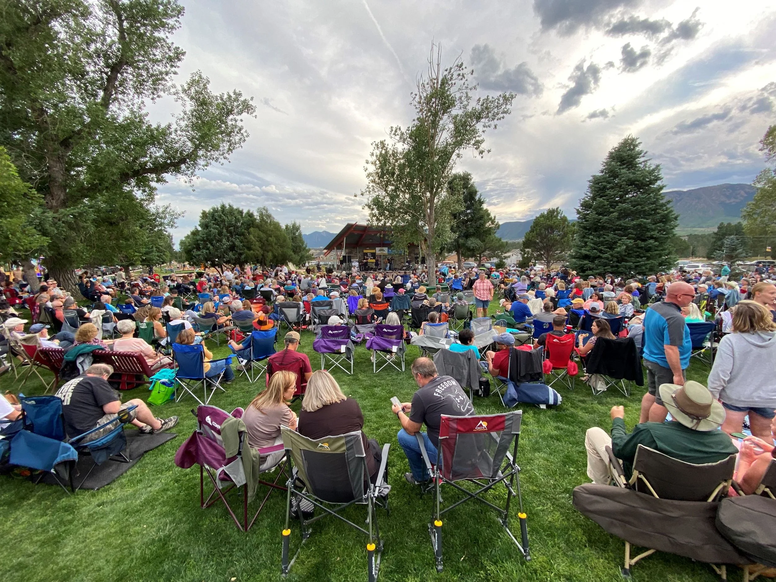 A large outdoor crowd gathered on a grassy area to watch a concert on a stage under a pavilion with mountains in the background. Many people are seated in lawn chairs and some are standing, with trees and cloudy sky in the scene.
