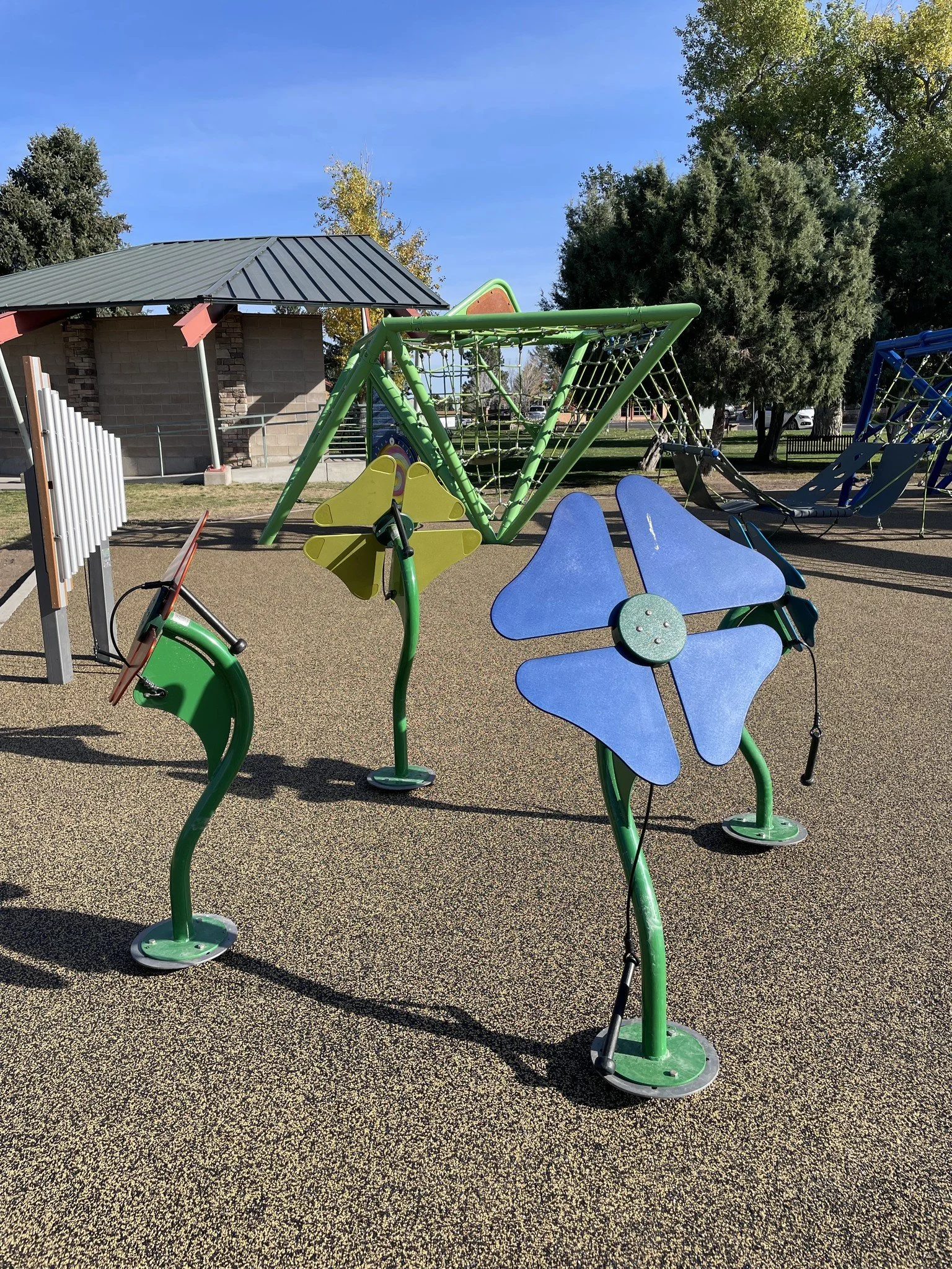 Colorful outdoor playground equipment with blue flower-shaped panels, green curved poles, and climbing structures, set on a rubberized surface in a park with trees and a pavilion under a clear blue sky.
