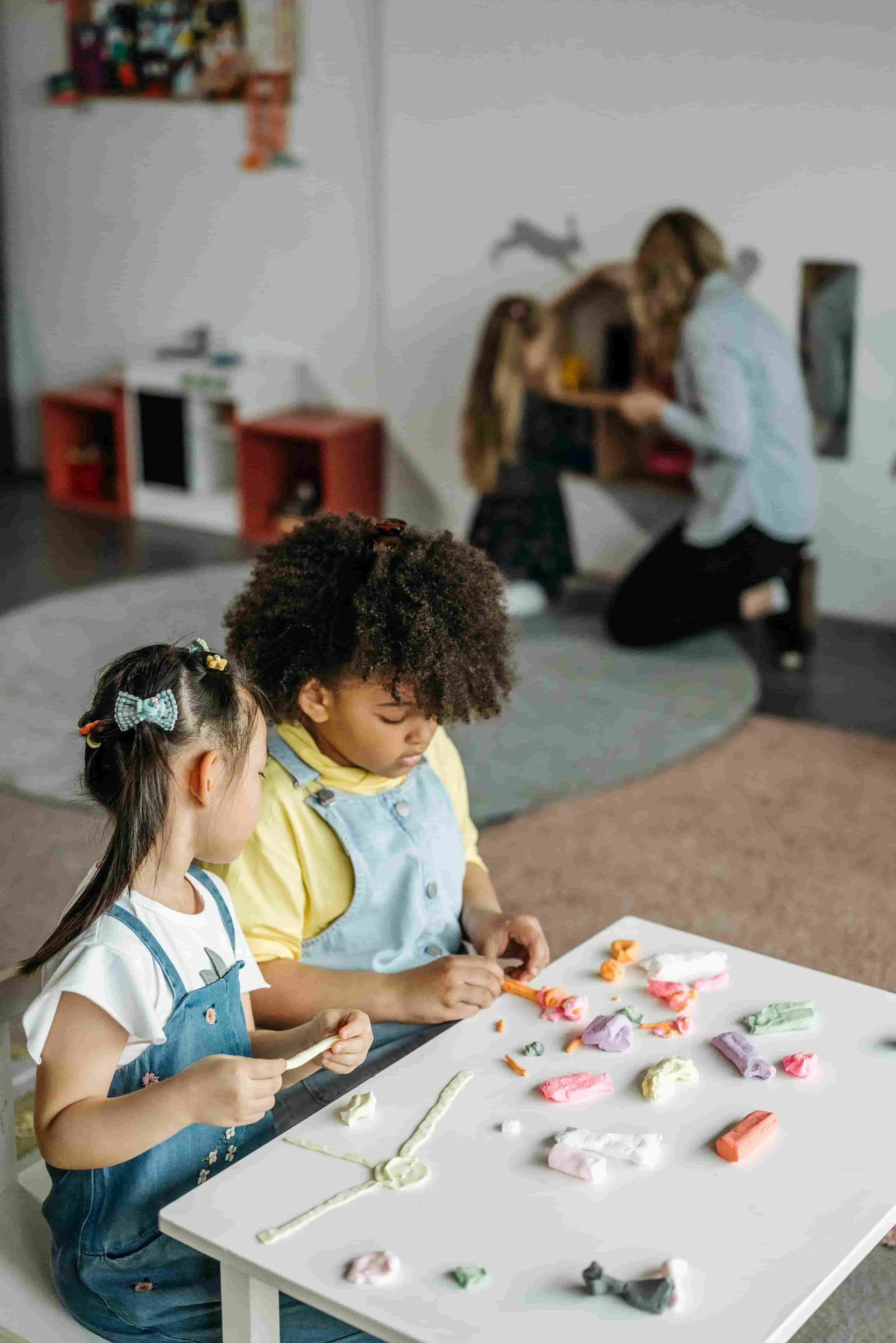 Three children sitting at a table making art with colorful modeling clay, with an adult assisting in the background.
