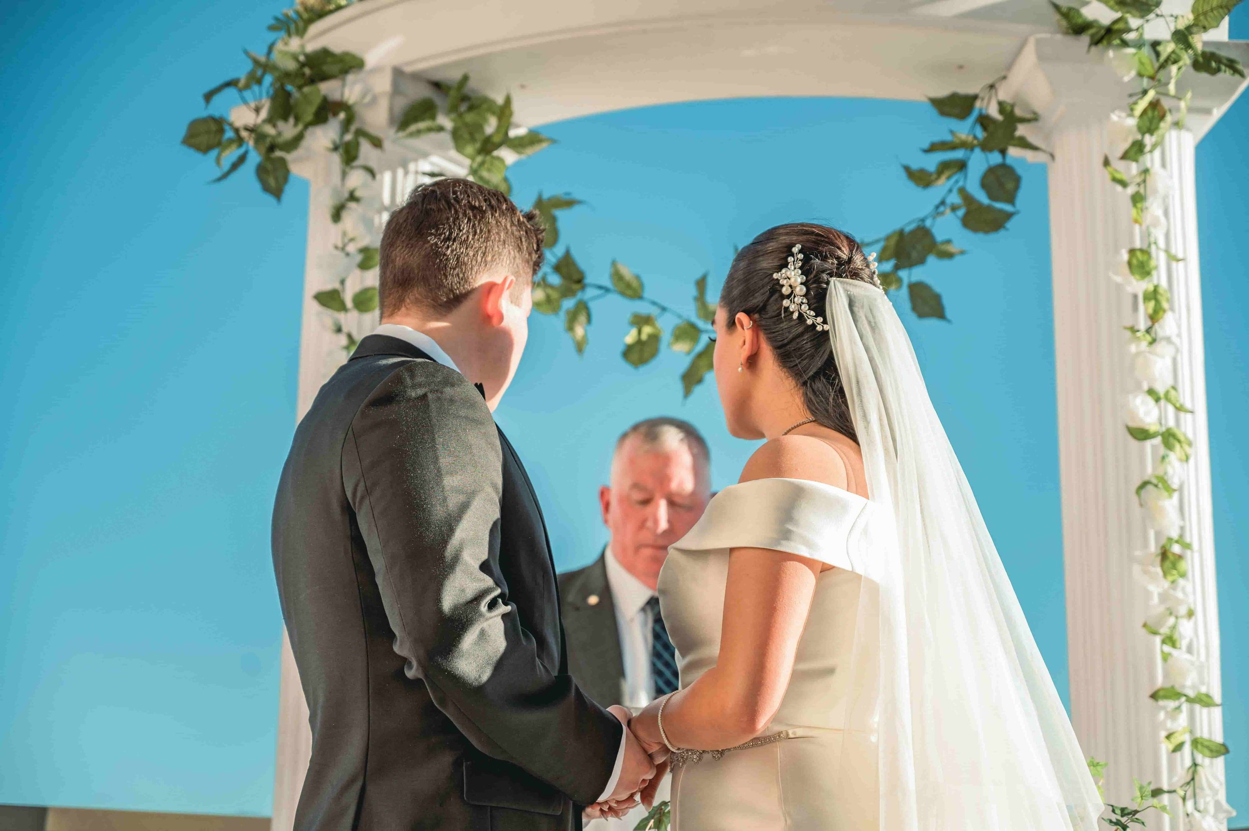 A bride and groom at their wedding ceremony, facing each other and holding hands, with an officiant standing behind them. The background includes a white arch decorated with green leaves and flowers, set against a bright blue sky.