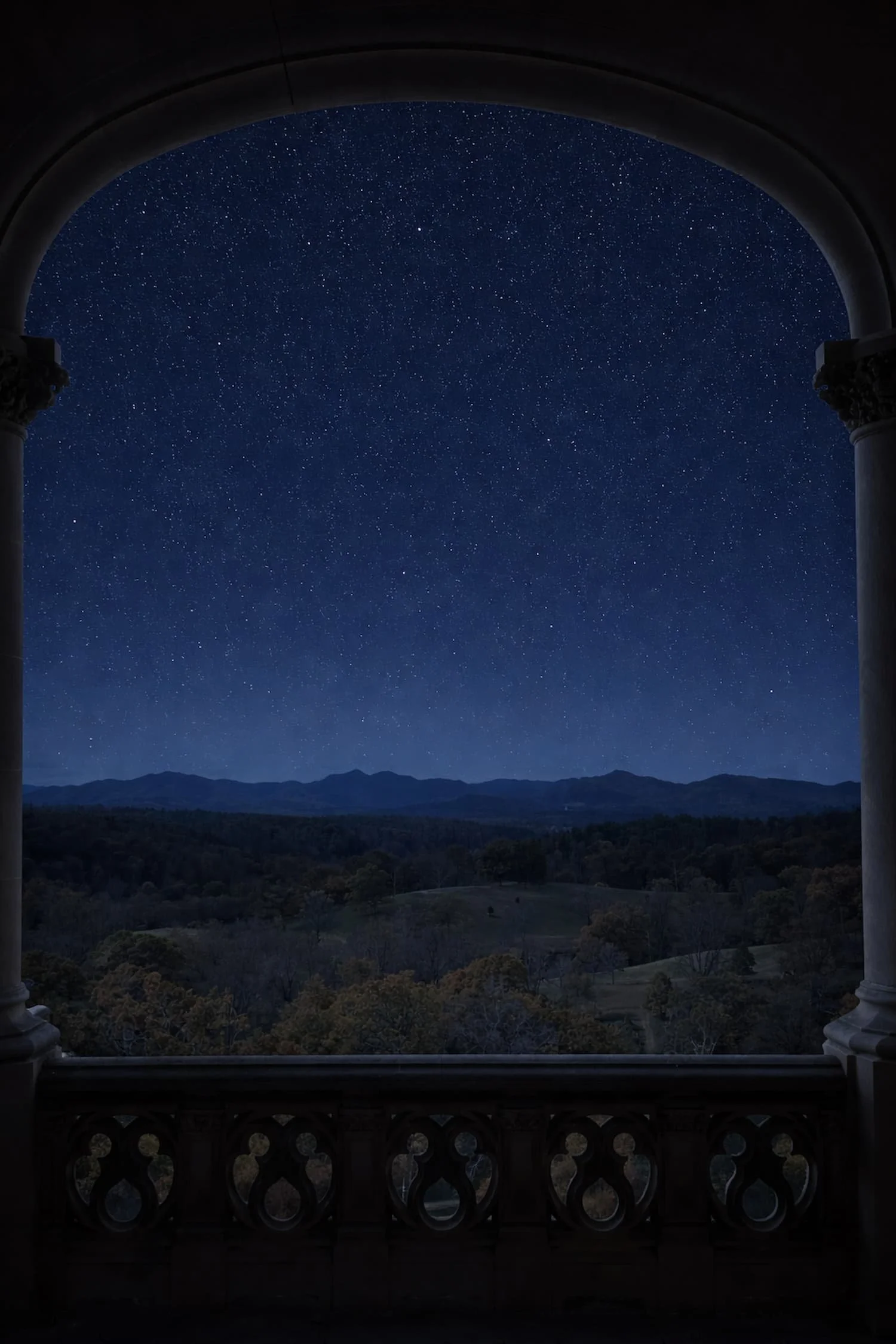 Night sky filled with stars visible through an arched balcony with decorative columns, overlooking a landscape of rolling hills and trees located at the Biltmore Estate in Asheville, NC.