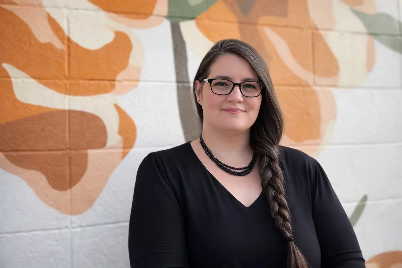 Kelly Goulet, A woman with dark brown hair styled in a braid, wearing glasses, a black top, and a black beaded necklace, standing in front of a colorful mural of Flowers with light orange and beige leaves on a white background.
