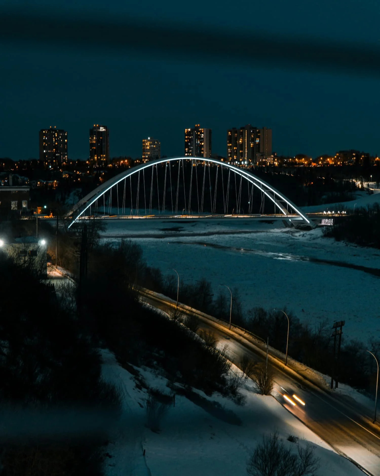 Night view of a Edmonton, AB with illuminated buildings and a lit modern arch bridge over a snow-covered area, with a winding road below.