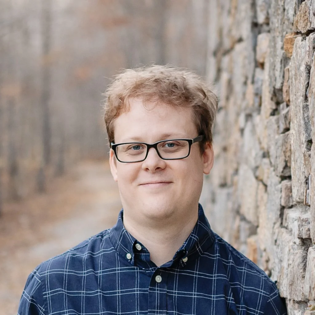 Tom Goulet, A man with light skin, glasses, and short, curly, light brown hair smiling in front of a rustic stone wall in an outdoor setting.