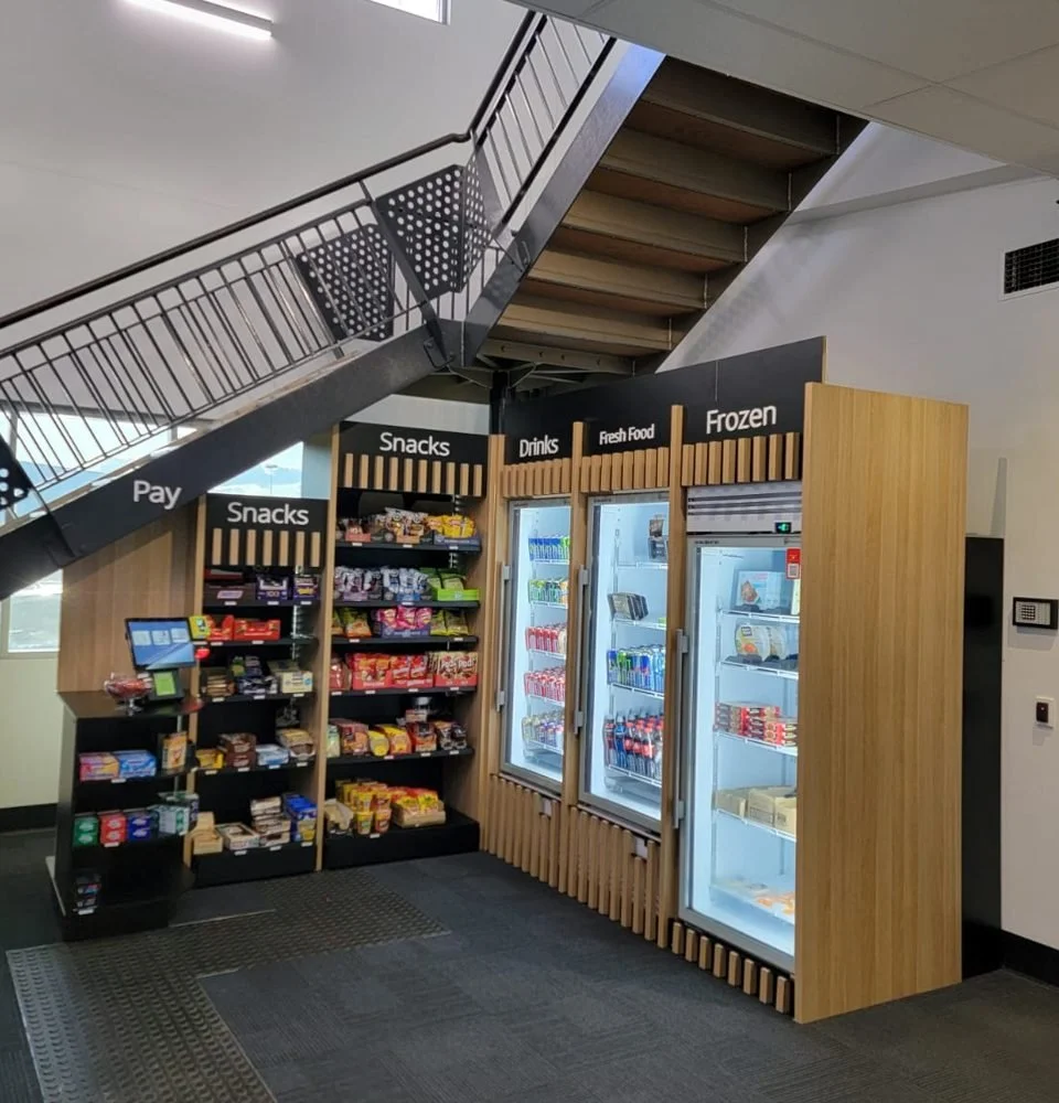 Workplace micro market with a self-serve payment station, snack shelving and drinks, fresh food and frozen fridges