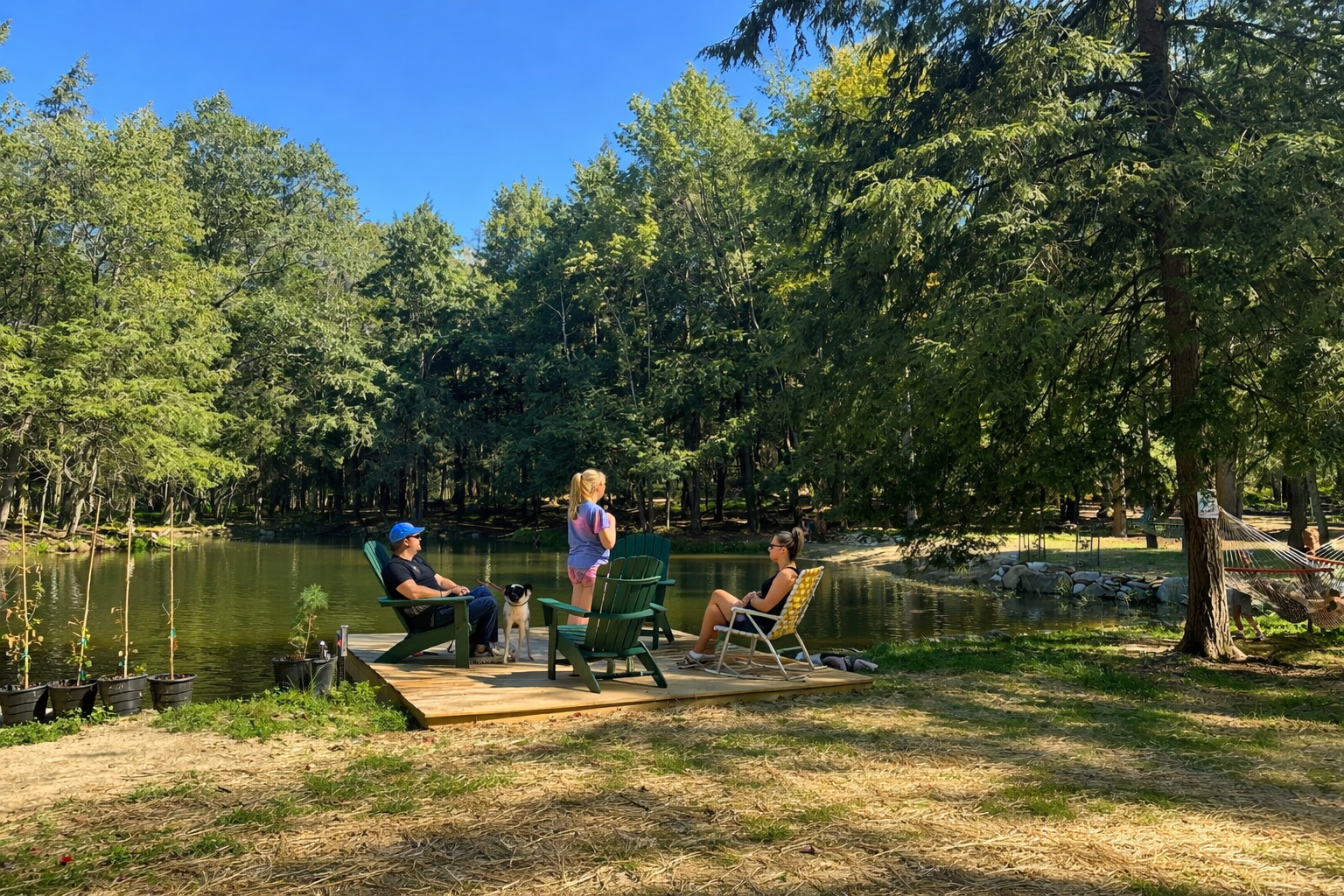 Three people and a dog sitting on a wooden dock by a lake, surrounded by trees, with one person standing and two seated on lawn chairs, sunny day with blue sky.