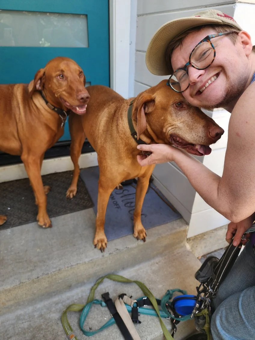 A man with glasses and a hat happily hugging two brown dogs in front of a blue door.