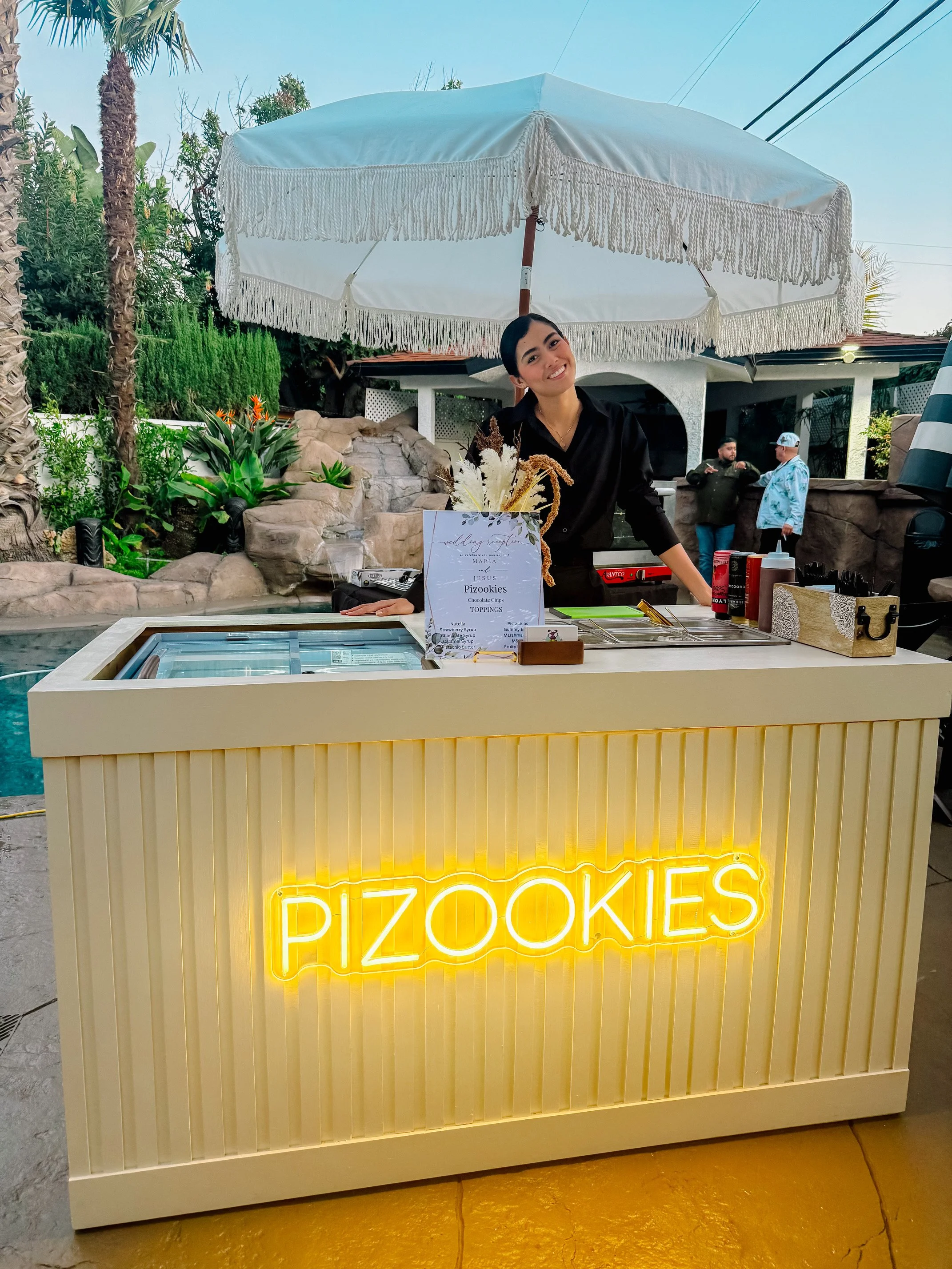 A person holding a bowl of ice cream with rainbow-colored candies and a white scoop of ice cream on top at an ice cream shop. The shop has various toppings in containers and a decorative vase with dried flowers on the counter.
