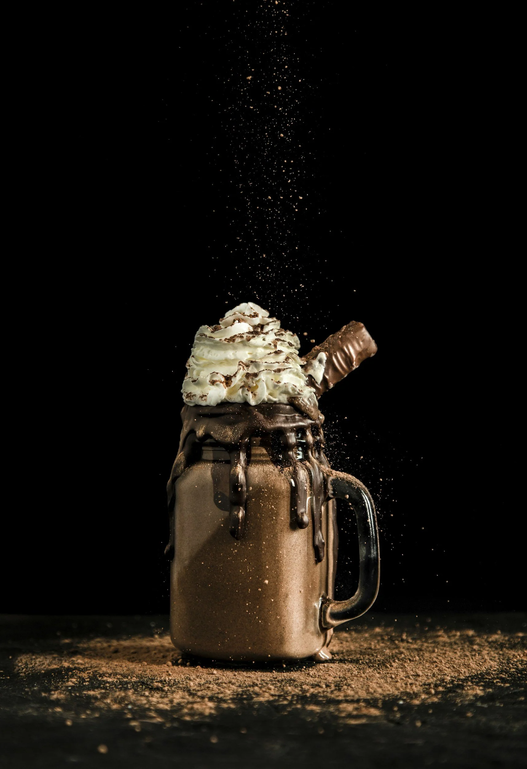A jar-style mug filled with a chocolate milkshake, topped with whipped cream, chocolate sprinkles, a chocolate bar, and caramel, with cocoa powder sprinkled on top and falling from above, set against a dark background.