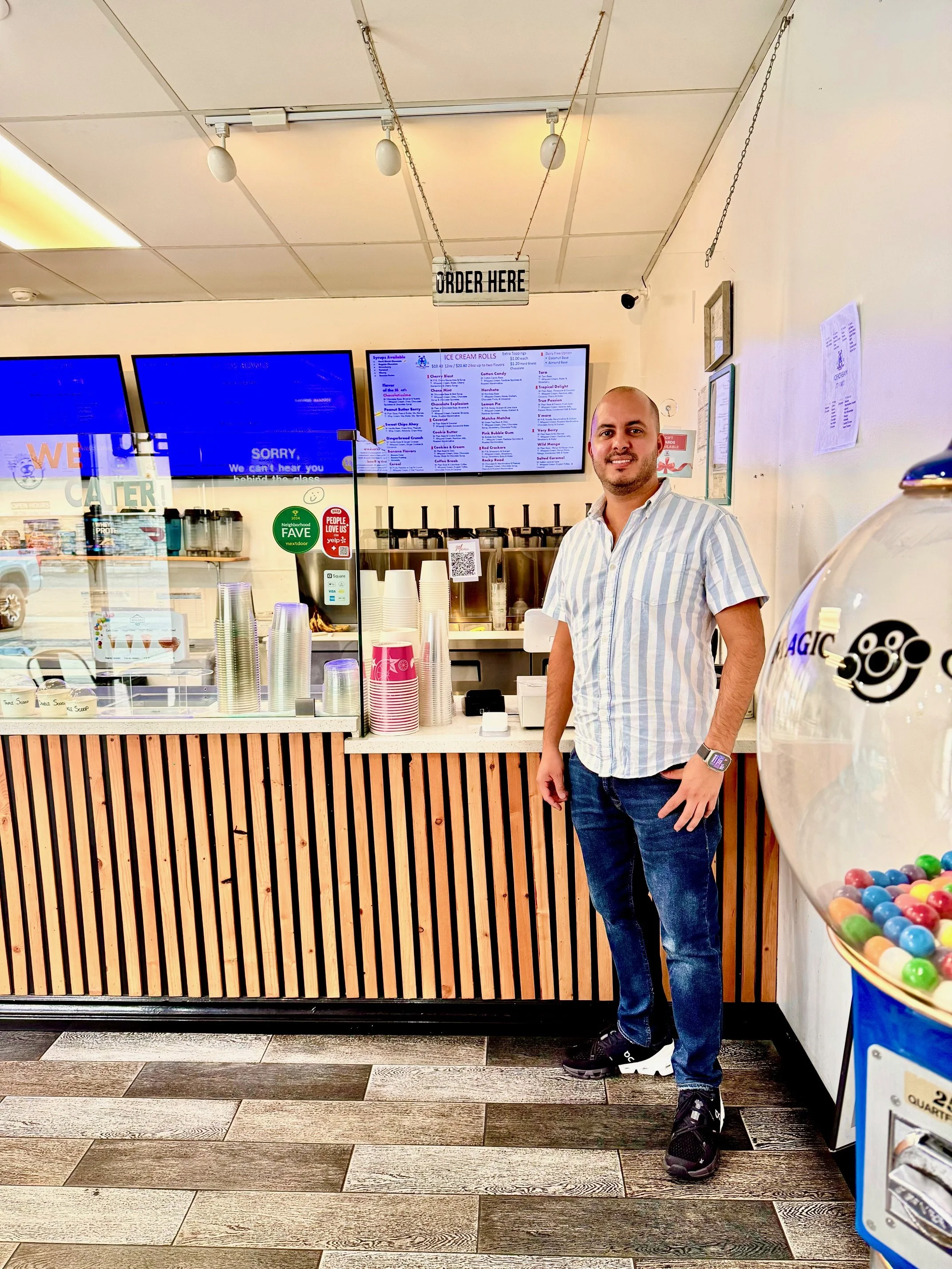 A man with a beard, wearing a striped short-sleeve shirt, jeans, and sneakers, standing inside a bubble tea shop next to a gumball machine, with a counter and digital menu screens in the background.