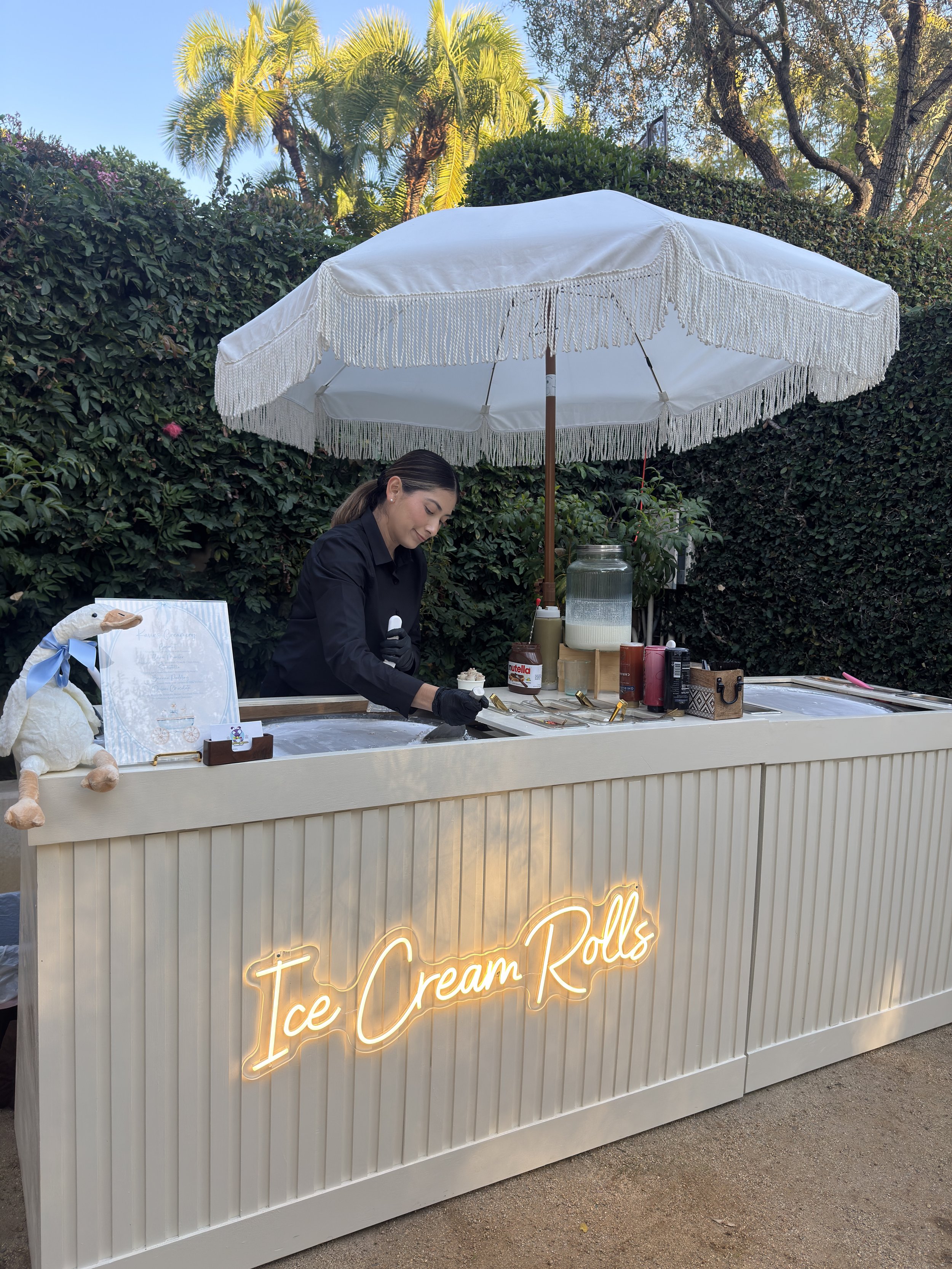 A woman working at an ice cream roll stand with a sign that reads 'Ice Cream Rolls'. The stand has a white canopy with fringe, and is set outdoors with trees and palm trees in the background.