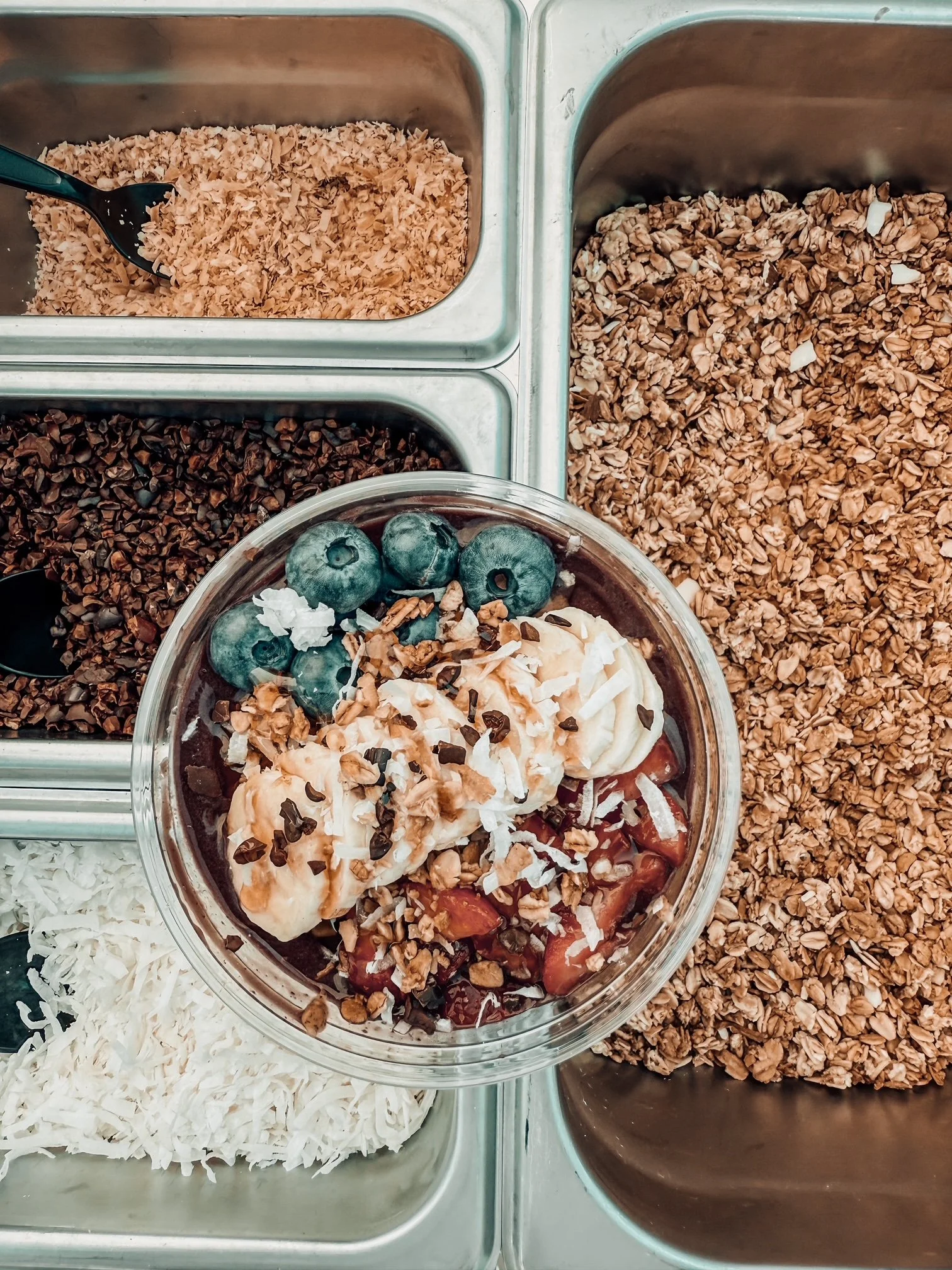 Various toppings and ingredients for a frozen yogurt or ice cream parfait, including blueberries, shredded coconut, chopped nuts, chocolate chips, and chopped cookies, displayed in metal containers and a glass bowl.