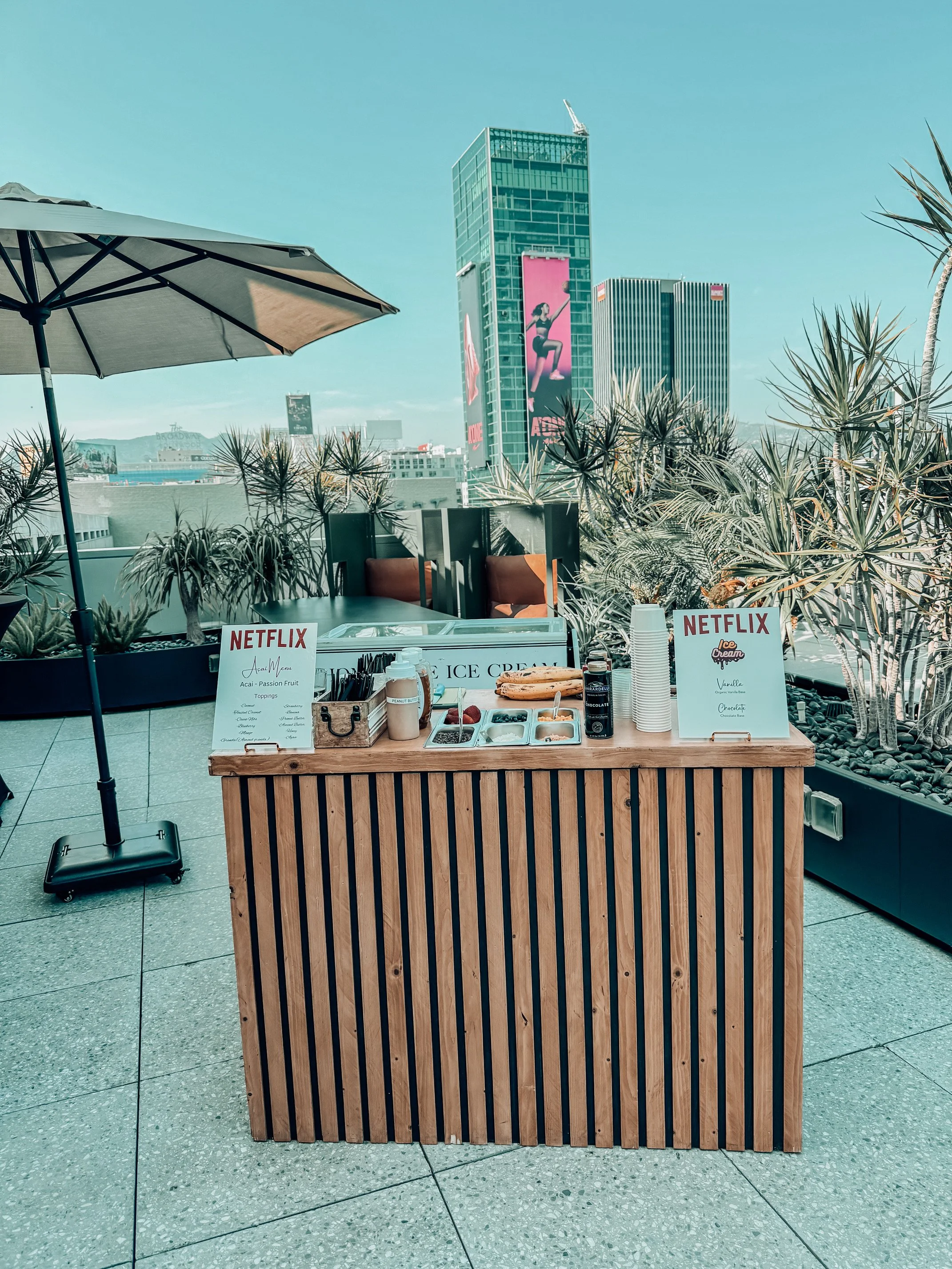Rooftop ice cream stand with toppings, signs promoting Netflix, umbrellas, and city skyline in the background.