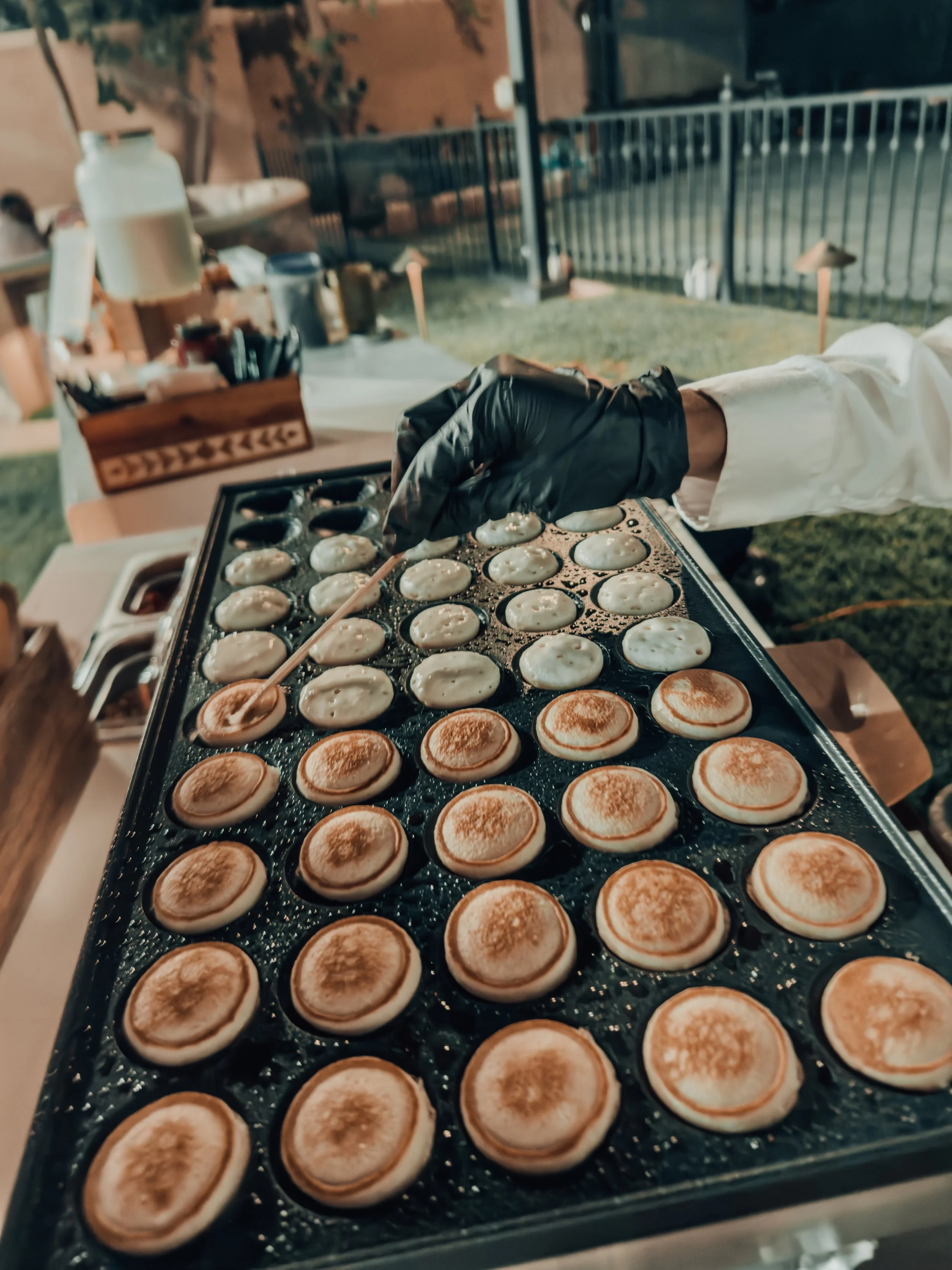 Person wearing a black glove cooking small round pancakes on a griddle during an outdoor event at night.