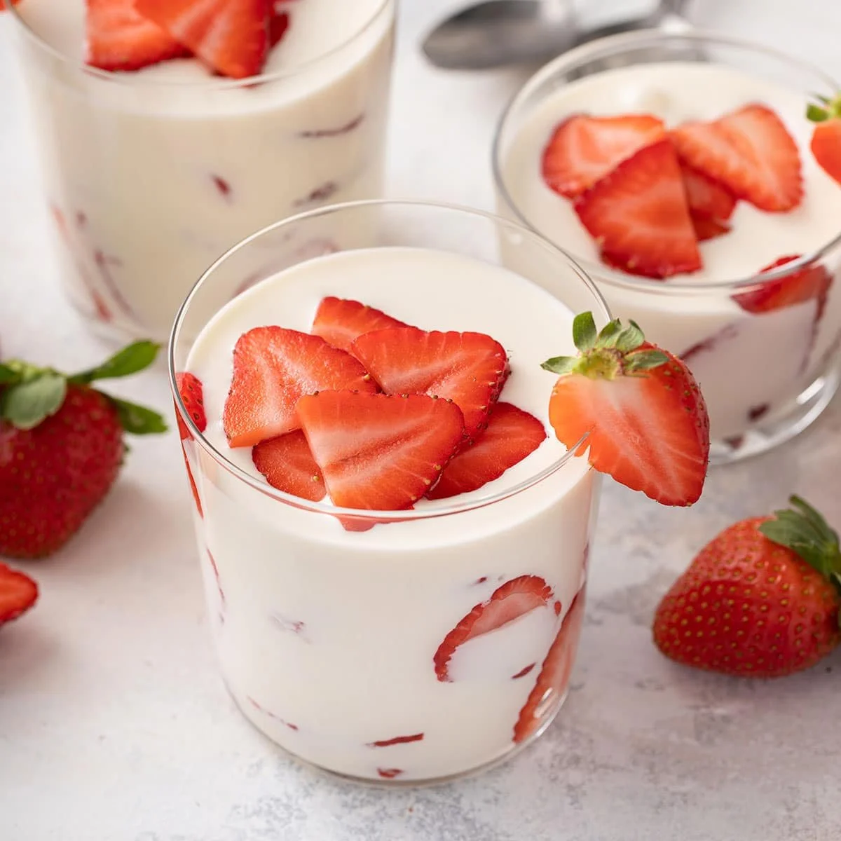 Glass cups filled with strawberry yogurt topped with sliced strawberries, some whole strawberries surrounding the cups.