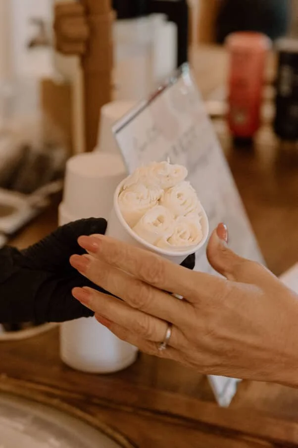 Two hands exchanging a small cup of vanilla ice cream, one hand wearing a black glove, the other with a wedding ring, in a cafe or ice cream shop.