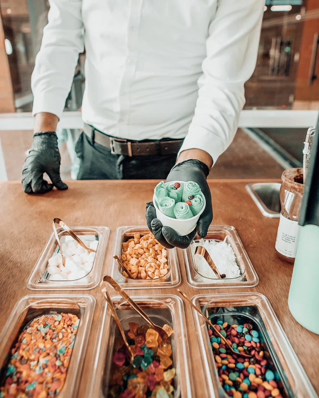 A woman making ice cream rolls at an outdoor stand with a neon sign that reads 'Ice Cream Rolls,' under a white parasol with fringe.