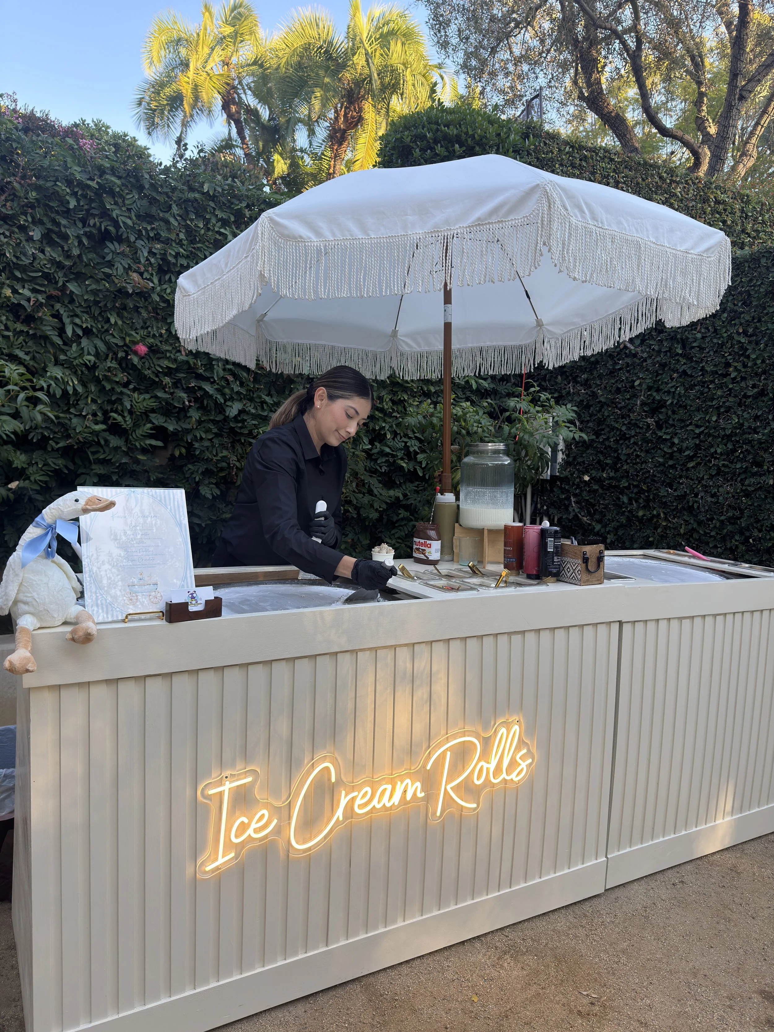 A woman working at an ice cream roll stand labeled "Ice Cream Rolls" with a white parasol, surrounded by greenery and palm trees.