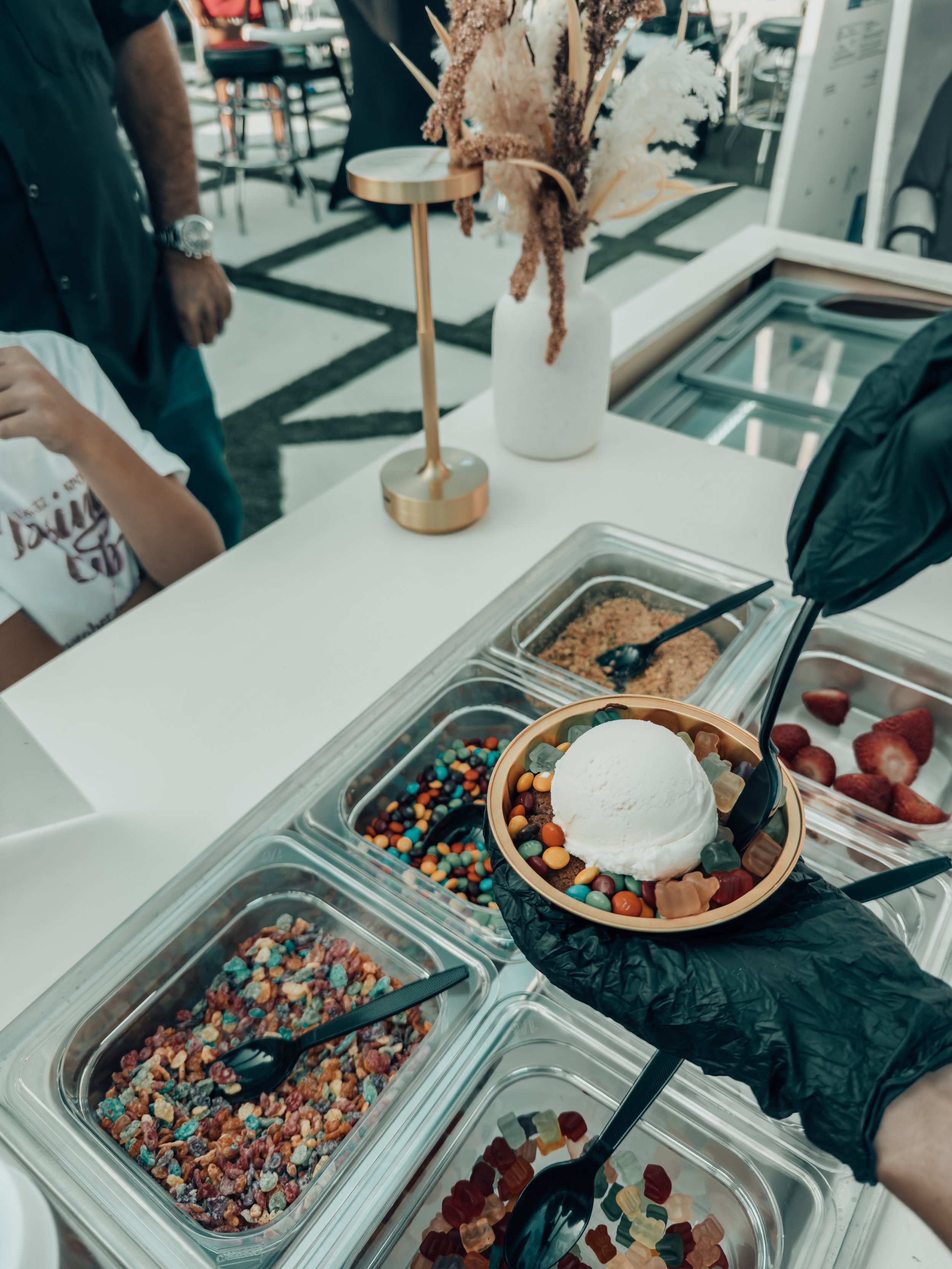 A person holding a bowl of ice cream with rainbow-colored candies and a white scoop of ice cream on top at an ice cream shop. The shop has various toppings in containers and a decorative vase with dried flowers on the counter.