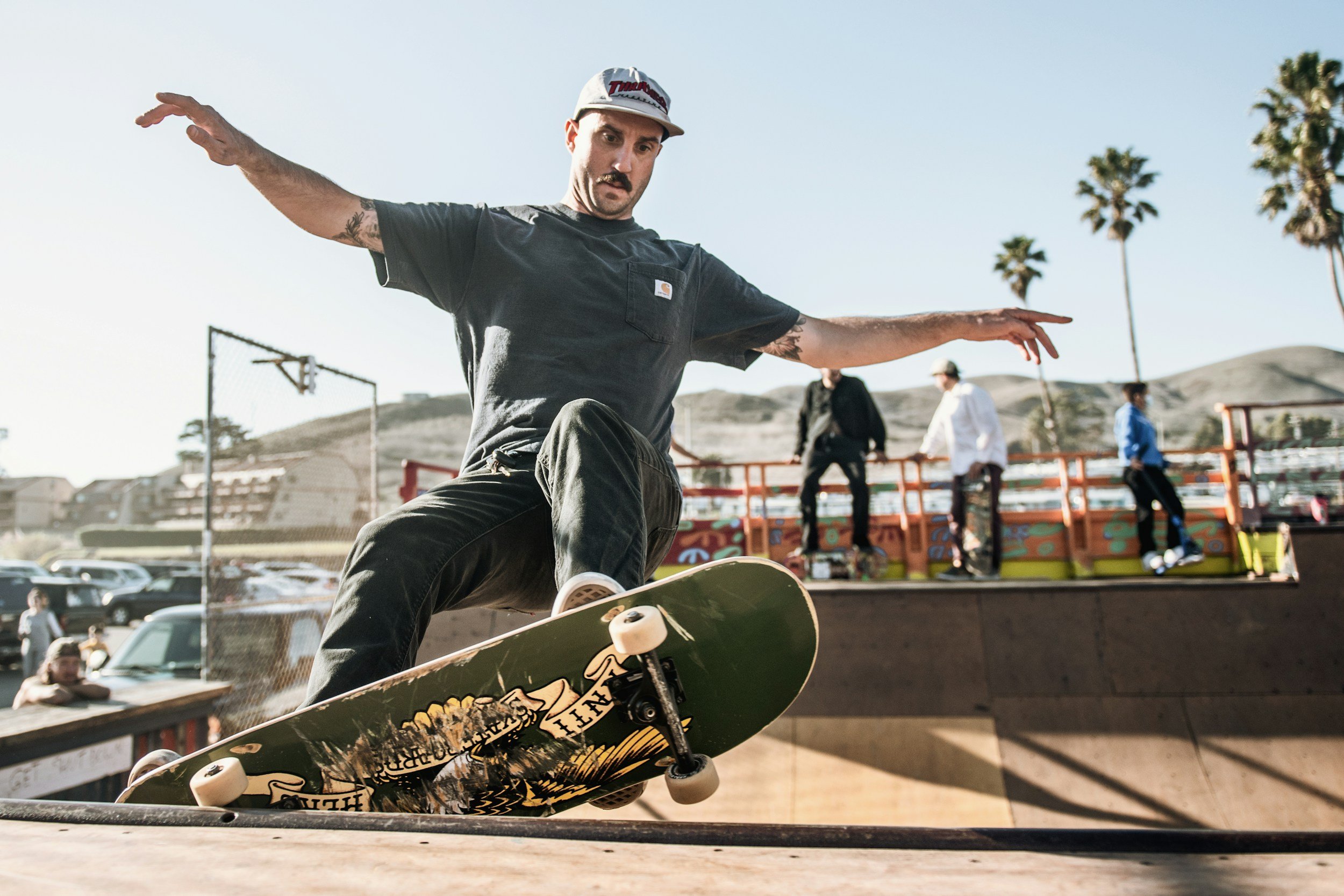 Skateboarder performing a trick at a skate park with onlookers and palm trees in the background.