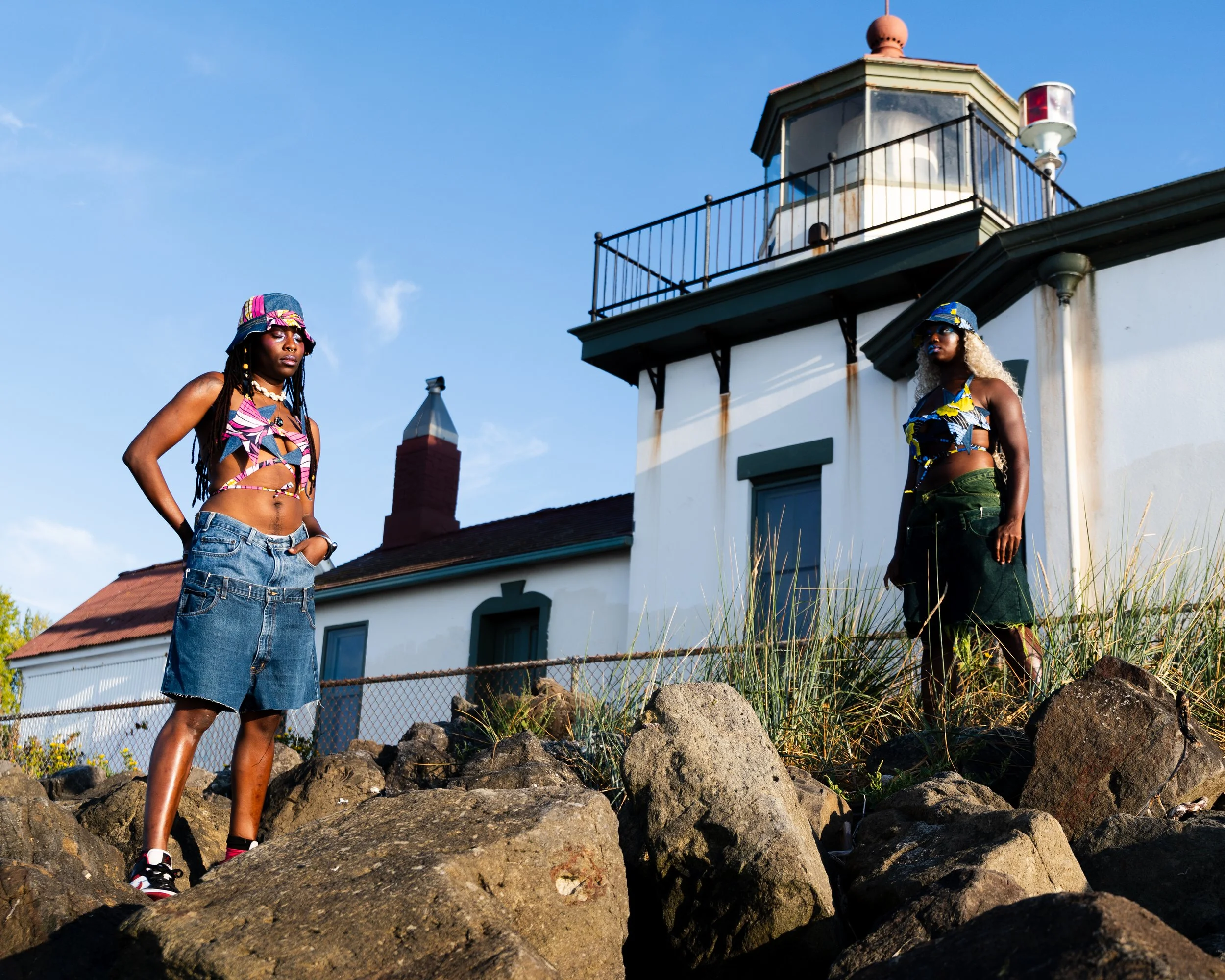 Two women with colorful hats and crop tops, one with dreadlocks and the other with curly blonde hair, stand on large rocks in front of a white lighthouse building on a sunny day.