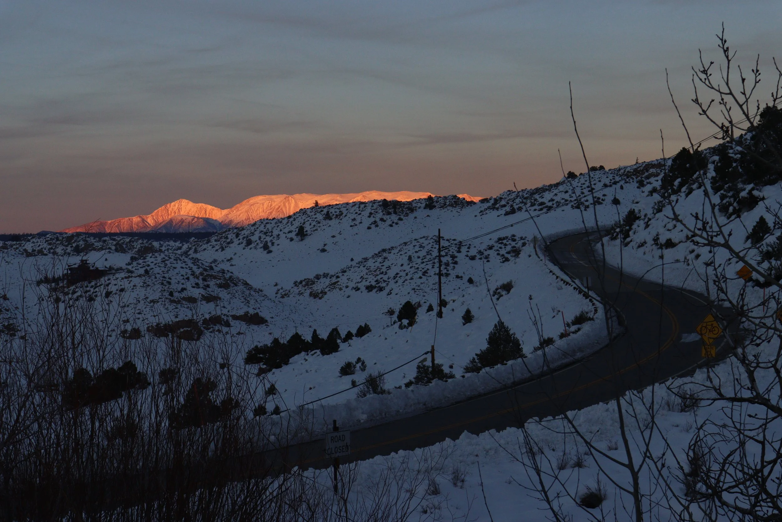 Snow-covered mountains at sunset with orange glow on peaks, winding road blocked by signs, snow-covered hills, and leafless trees in foreground.