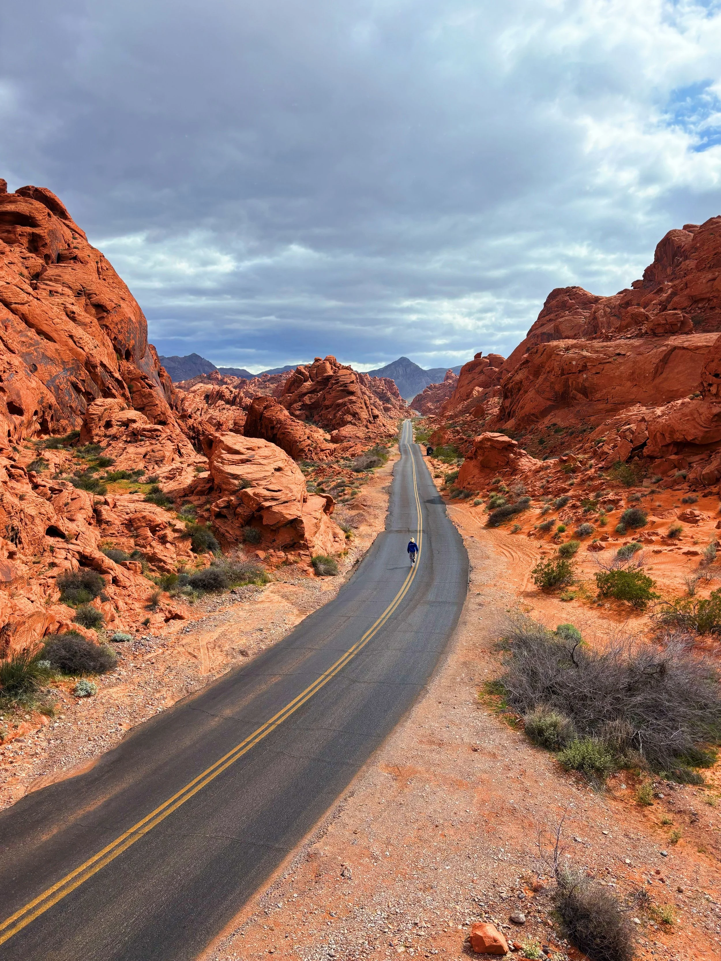 A long, straight road runs through a desert landscape with red rock formations on both sides, under a partly cloudy sky with some blue showing.