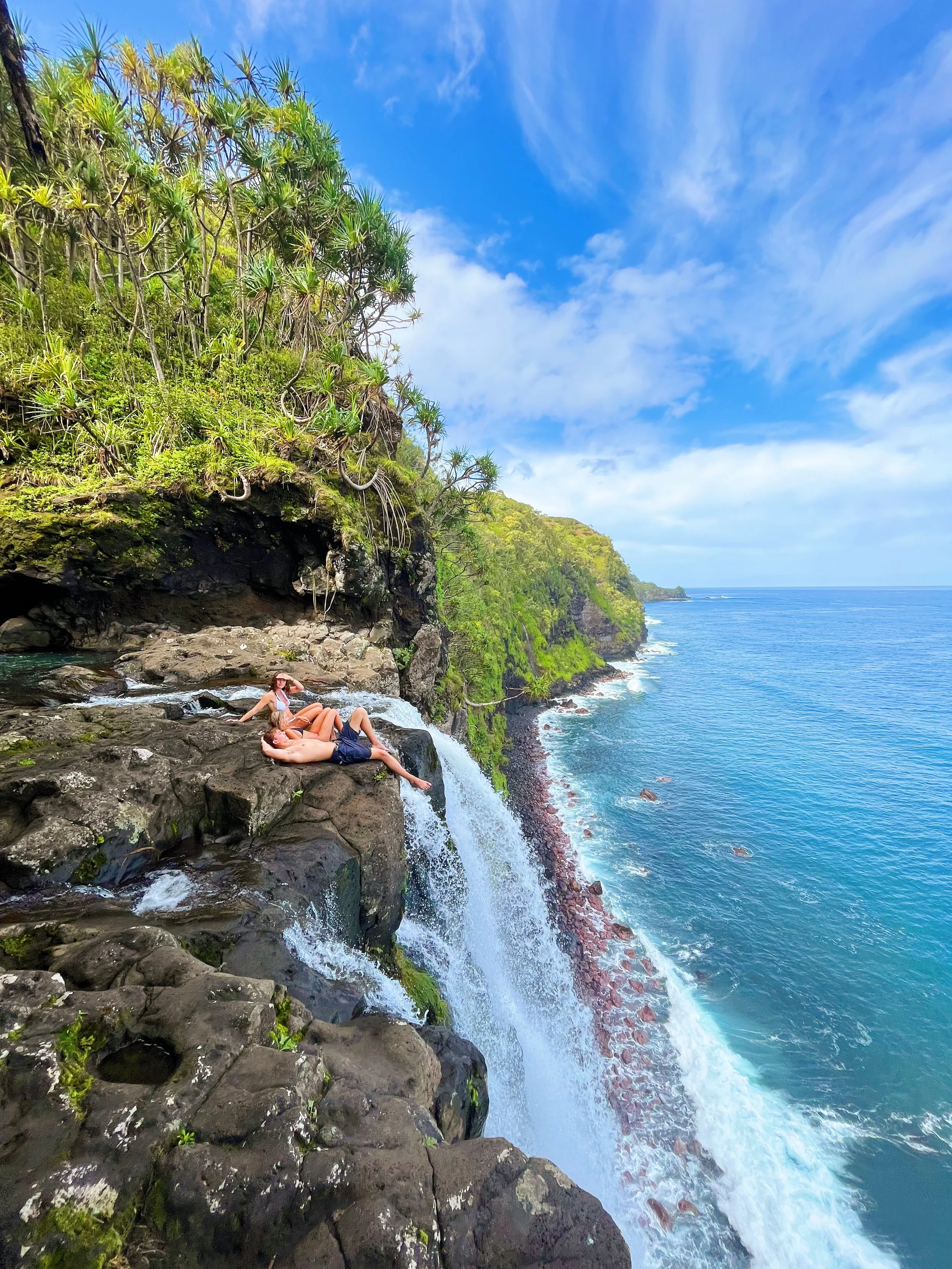 A couple relaxing on dark rocks near a small waterfall along a rugged coast with lush green cliffs and blue ocean under a partly cloudy sky.
