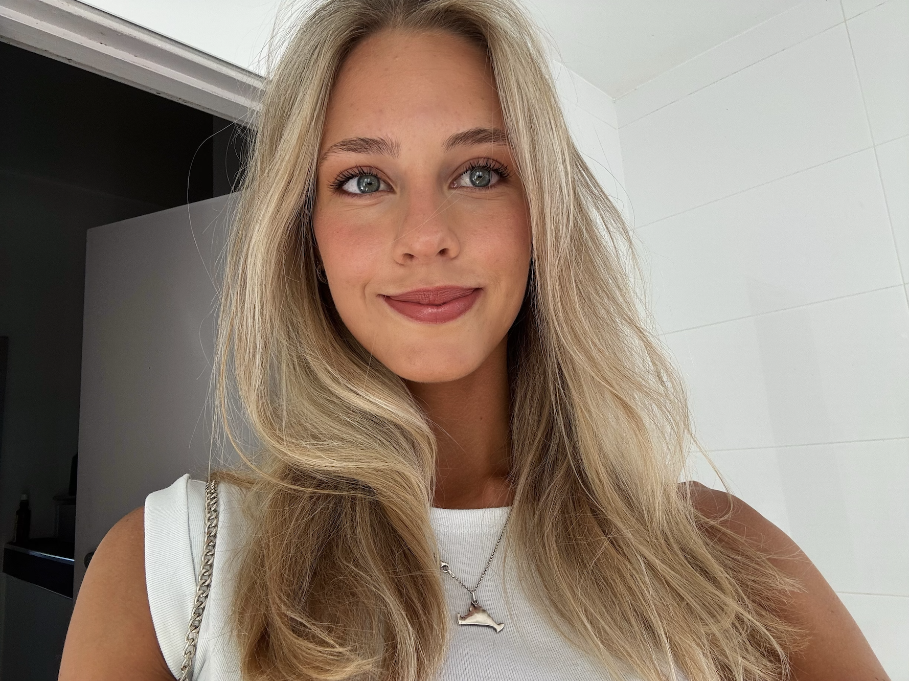 A young woman with long blonde hair, blue eyes, and light makeup, smiling at the camera in a well-lit room with white tiled walls.