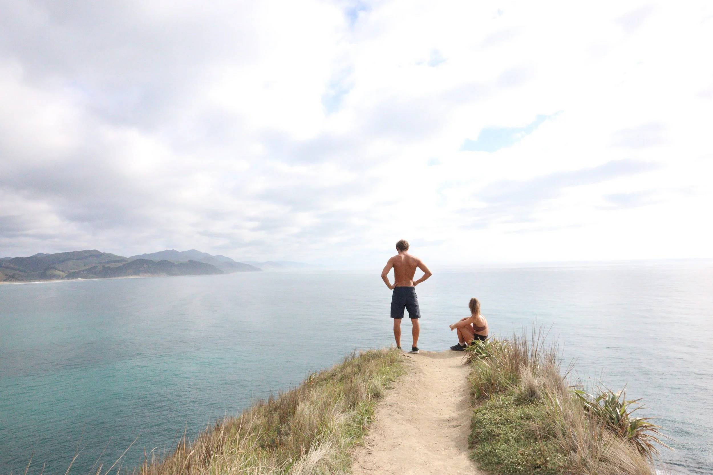 A man and woman overlook the ocean from a grassy cliffside path under a cloudy sky with mountains in the background.